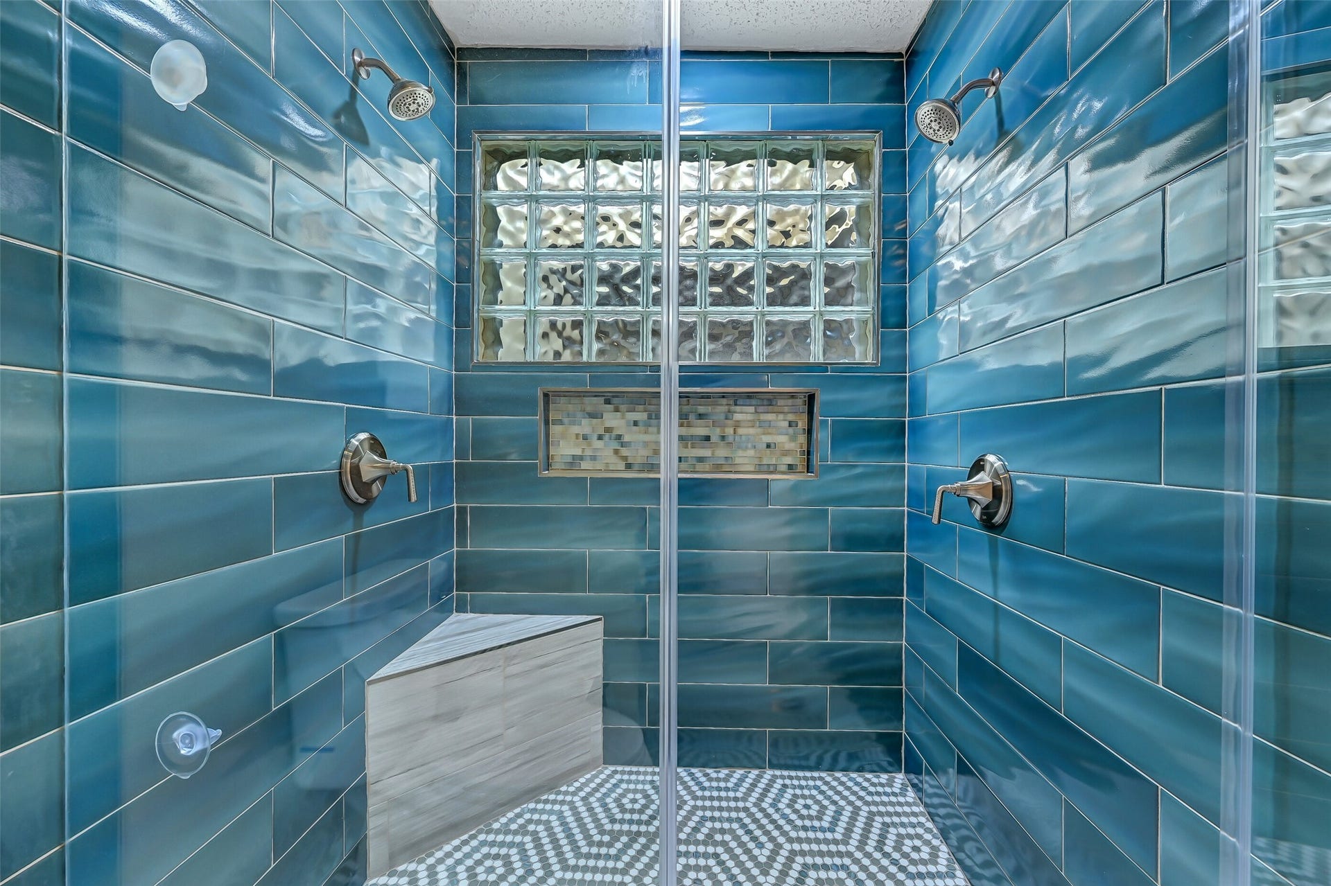 18214 Bal Harbour Drive, Nassau Bay, TX 77058 A striking feature of the remodeled primary bath is this spacious shower showcasing glossy blue subway tile with a toiletry niche, dual brushed-nickel showerheads, a corner bench, seamless shower glass doors, and a bright glass block window.