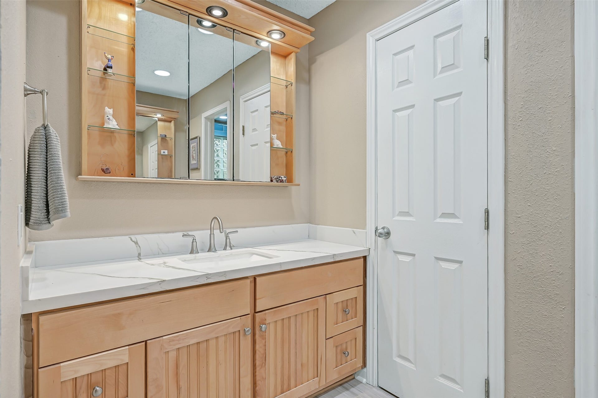 18214 Bal Harbour Drive, Nassau Bay, TX 77058 The remodeled primary bath features separate oak-finish vanities with quartz countertops, porcelain sinks & brushed-nickel fixtures. This vanity offers generous cabinet and drawer storage, plus a custom mirrored storage cabinet with glass display shelves & integrated LED lighting. A walk-in closet is located just beyond with attic access as well.