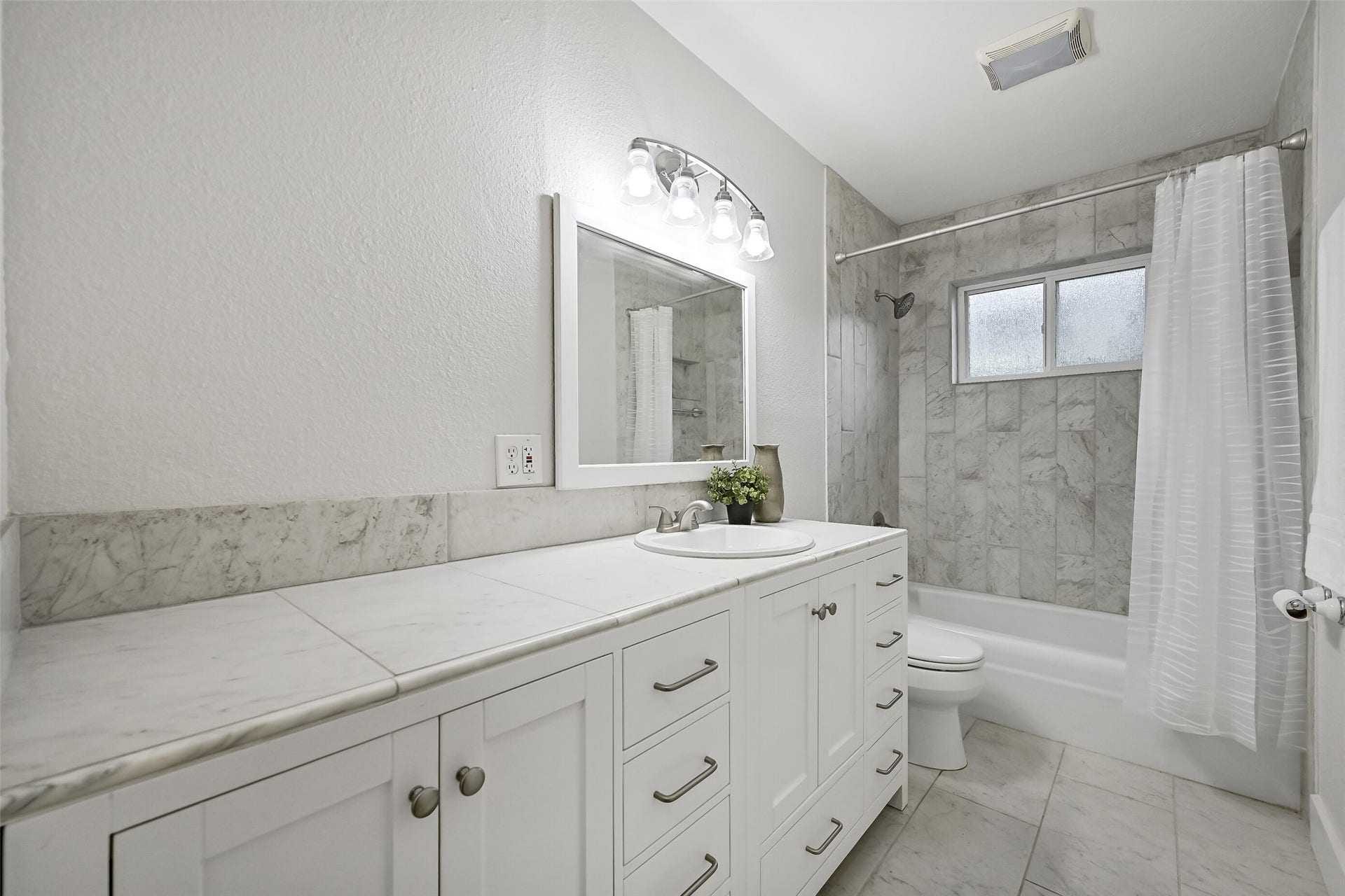 5403 Carmona Lane, Pearland, TX 77584 The updated guest bath features a long white vanity with plentiful soft-close drawer storage, large-format tile countertop, framed mirror & updated lighting. Tile flooring continues to the tub/shower combination, finished with a full-height marble-look tile surround & privacy window.