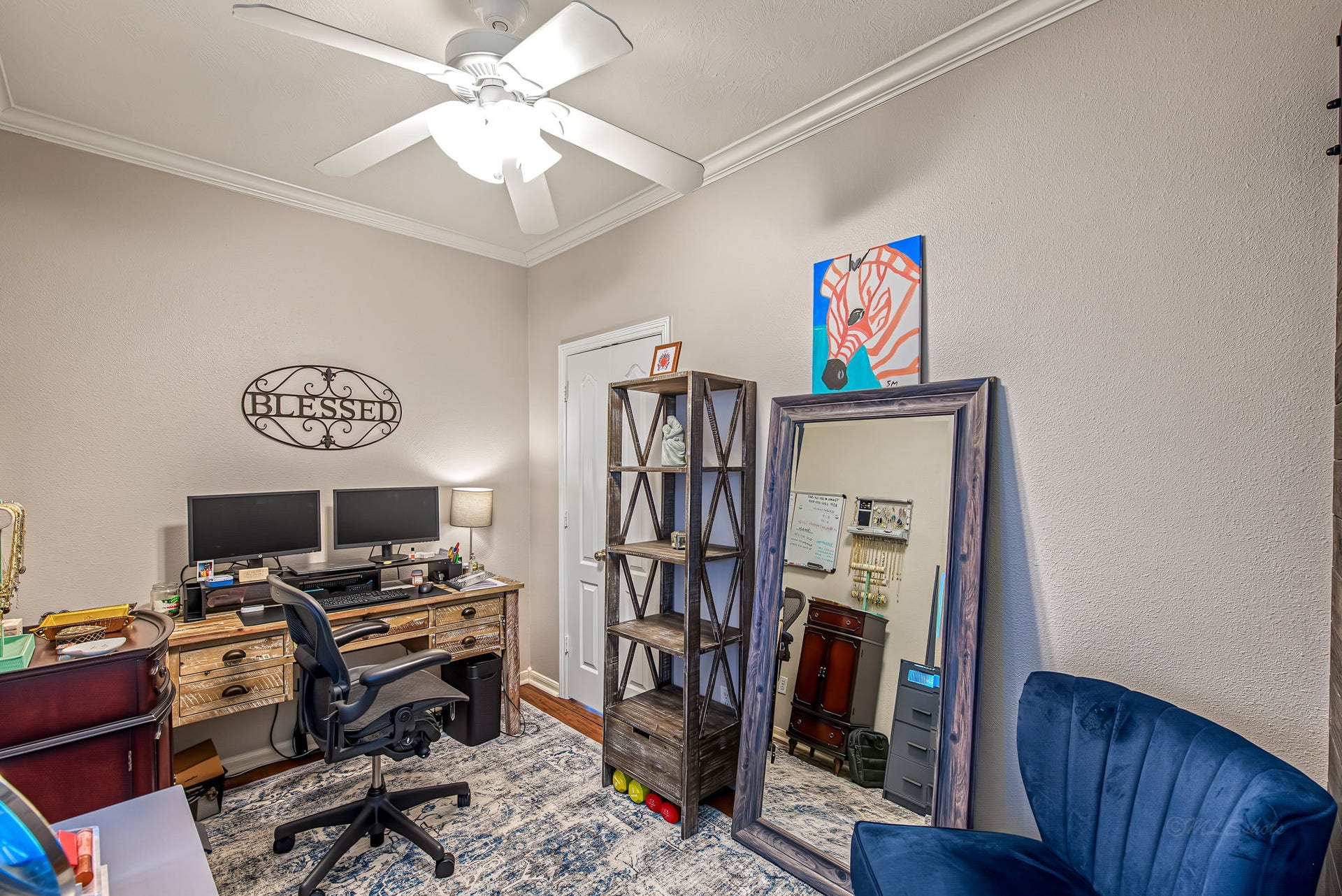 21223 Dover Park Lane, Katy, TX 77450 Charming home office featuring a spacious desk with dual monitors, wall shelving for storage, and a cozy blue chair, all illuminated by a ceiling fan light.