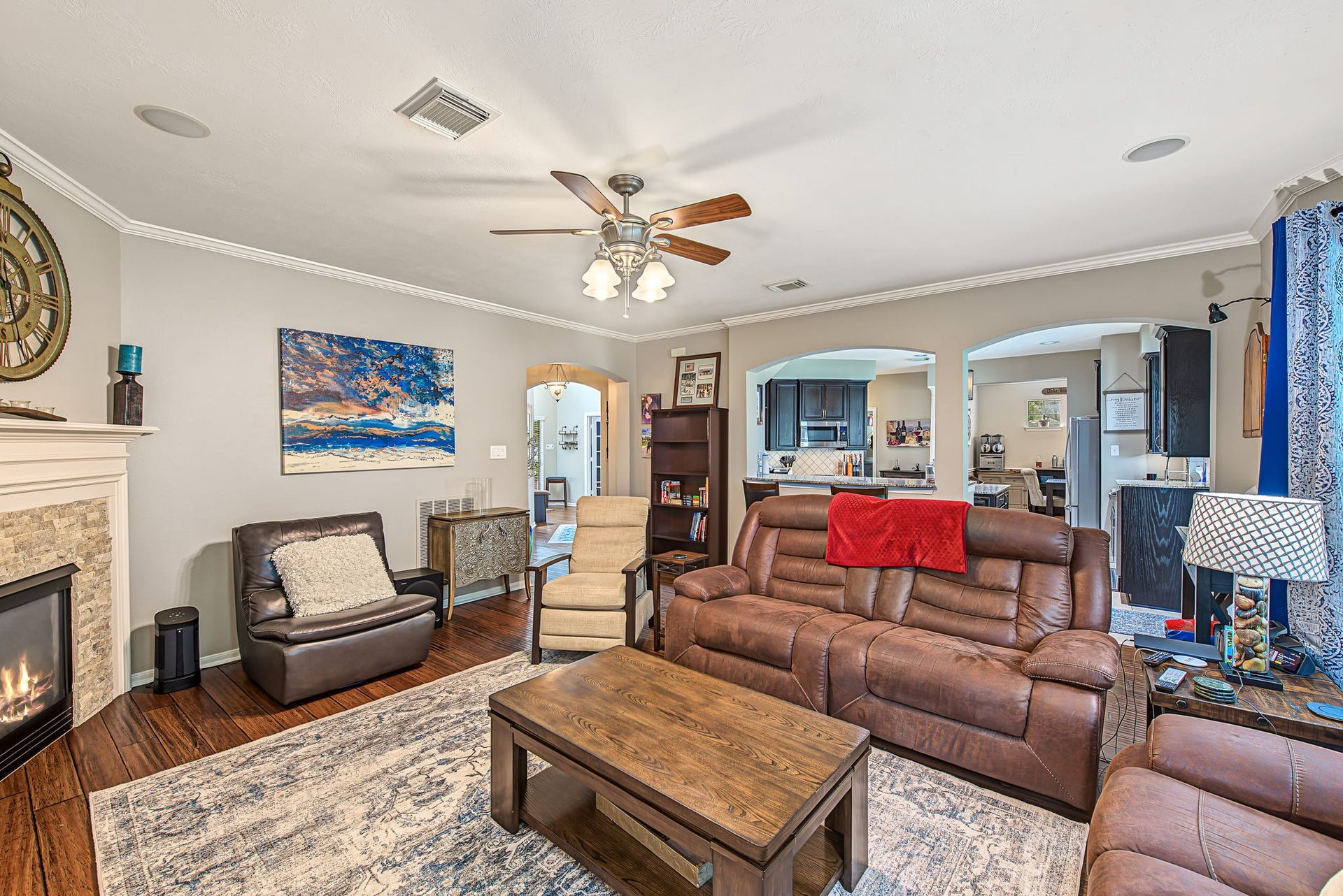21223 Dover Park Lane, Katy, TX 77450 Cozy living room featuring a stone fireplace, comfortable leather seating, and an elegant ceiling fan, seamlessly leading to the kitchen and dining areas through stylish archways.