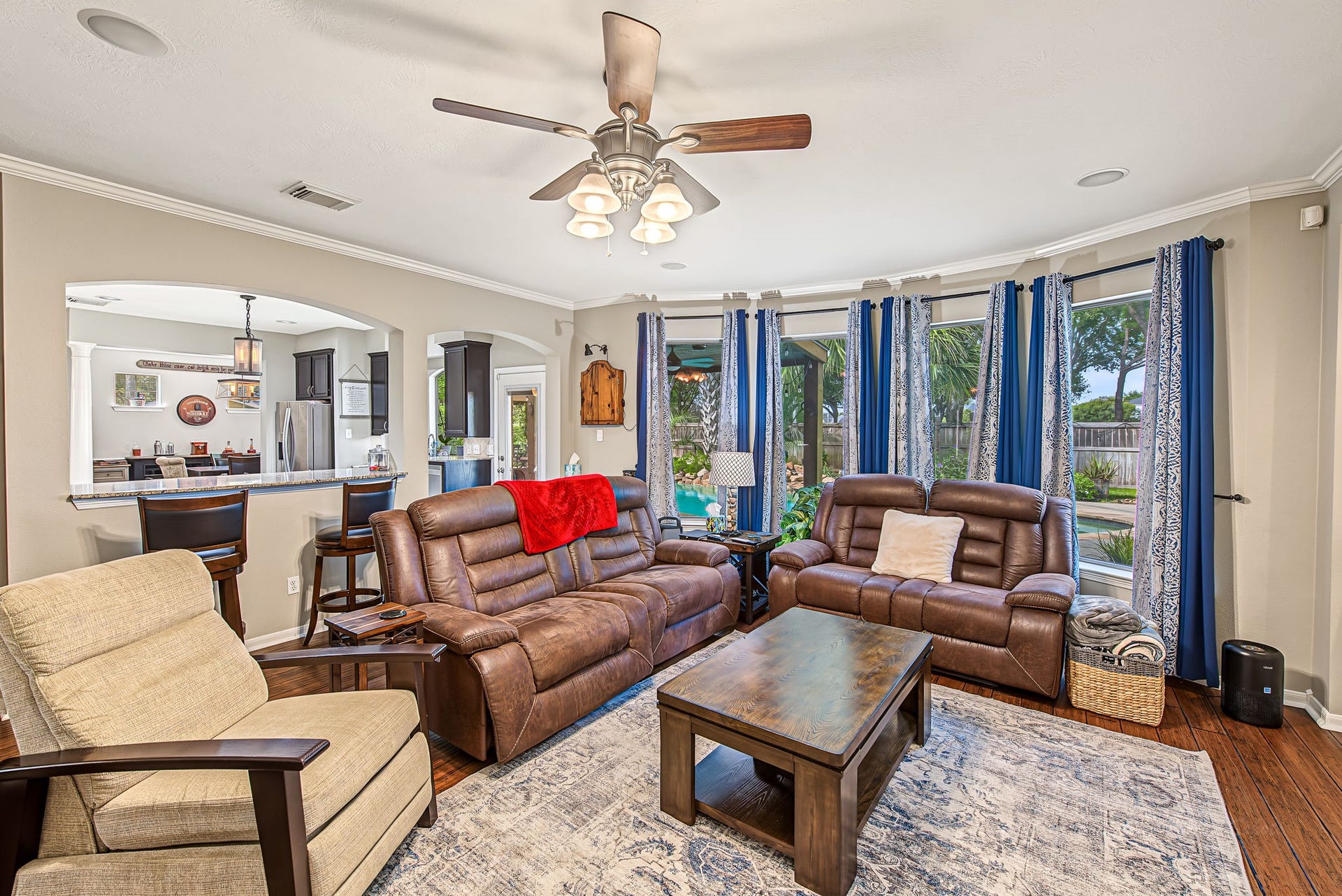 21223 Dover Park Lane, Katy, TX 77450 Bright and inviting living room featuring large windows with serene outdoor views, elegant hardwood floors, and comfortable leather seating, seamlessly connected to the kitchen area through a stylish open archway.