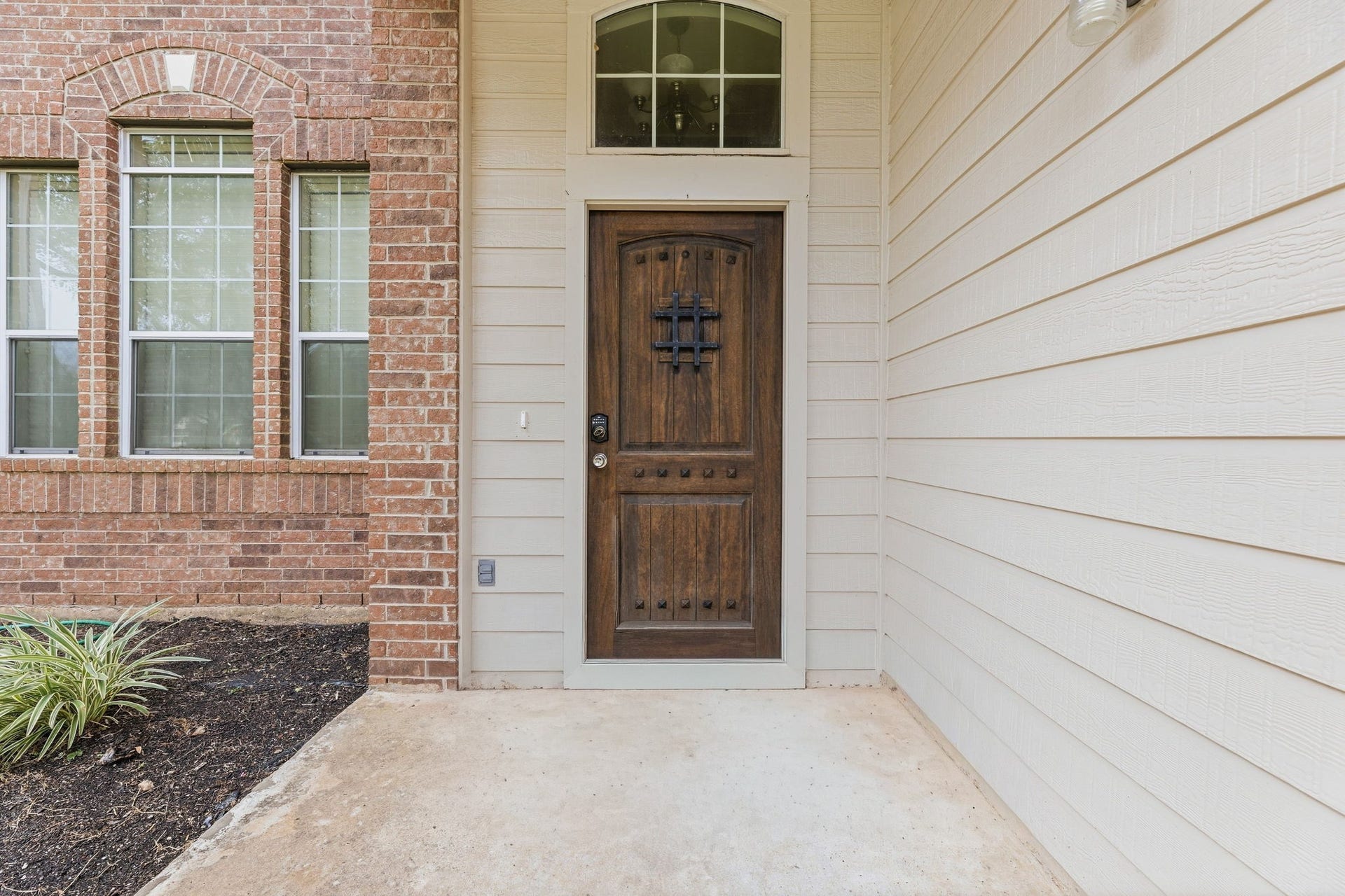 A covered brick archway frames a rustic wood front door with iron speakeasy detail, a transom window, and a keypad smart lock.