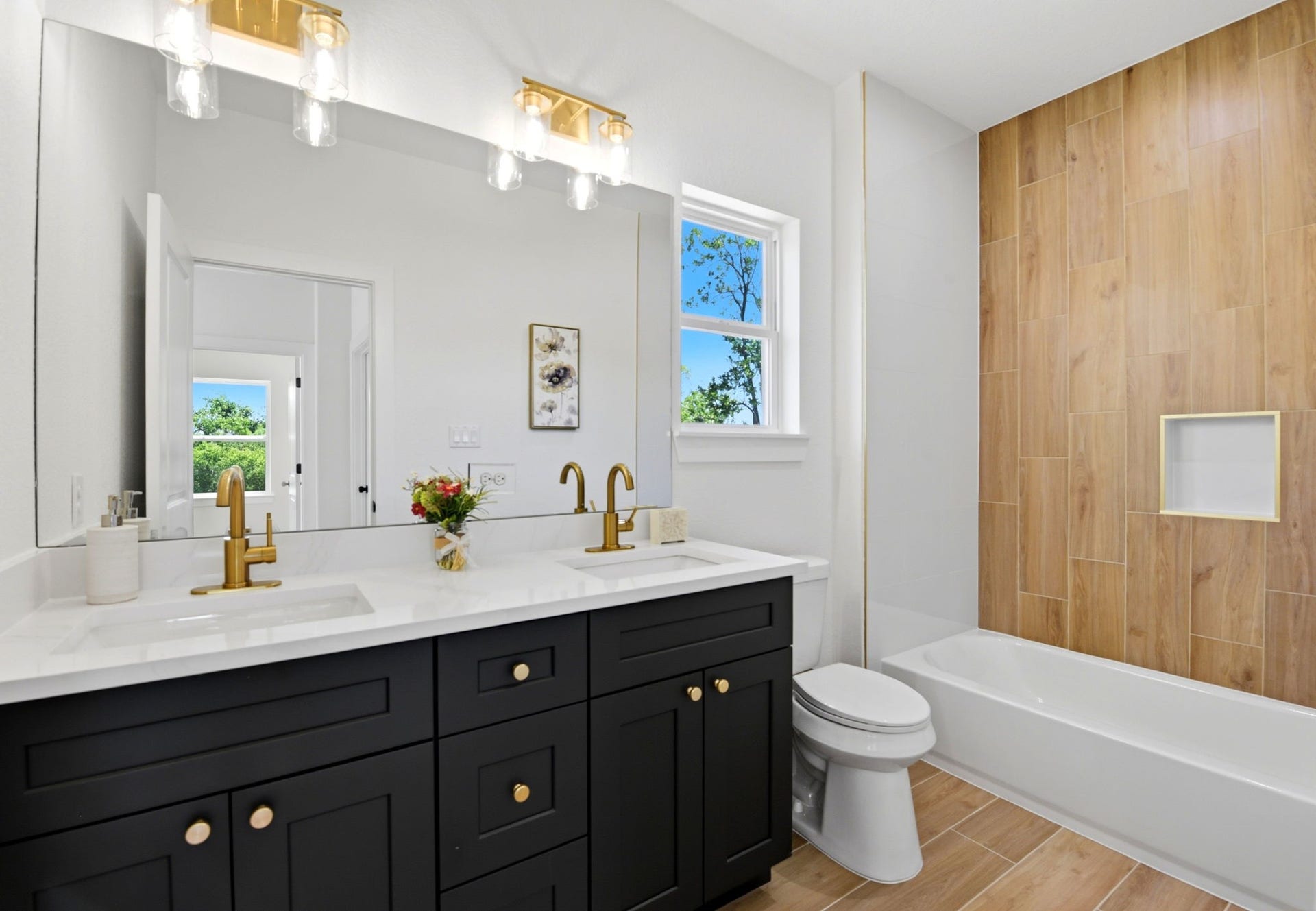 6117 Haight Street, Houston, TX 77028 Modern secondary bathroom with dual sinks and sleek black cabinetry. Features gold fixtures, a large mirror, and a window for natural light. The bathtub area has wood-style tiles for a warm touch.
