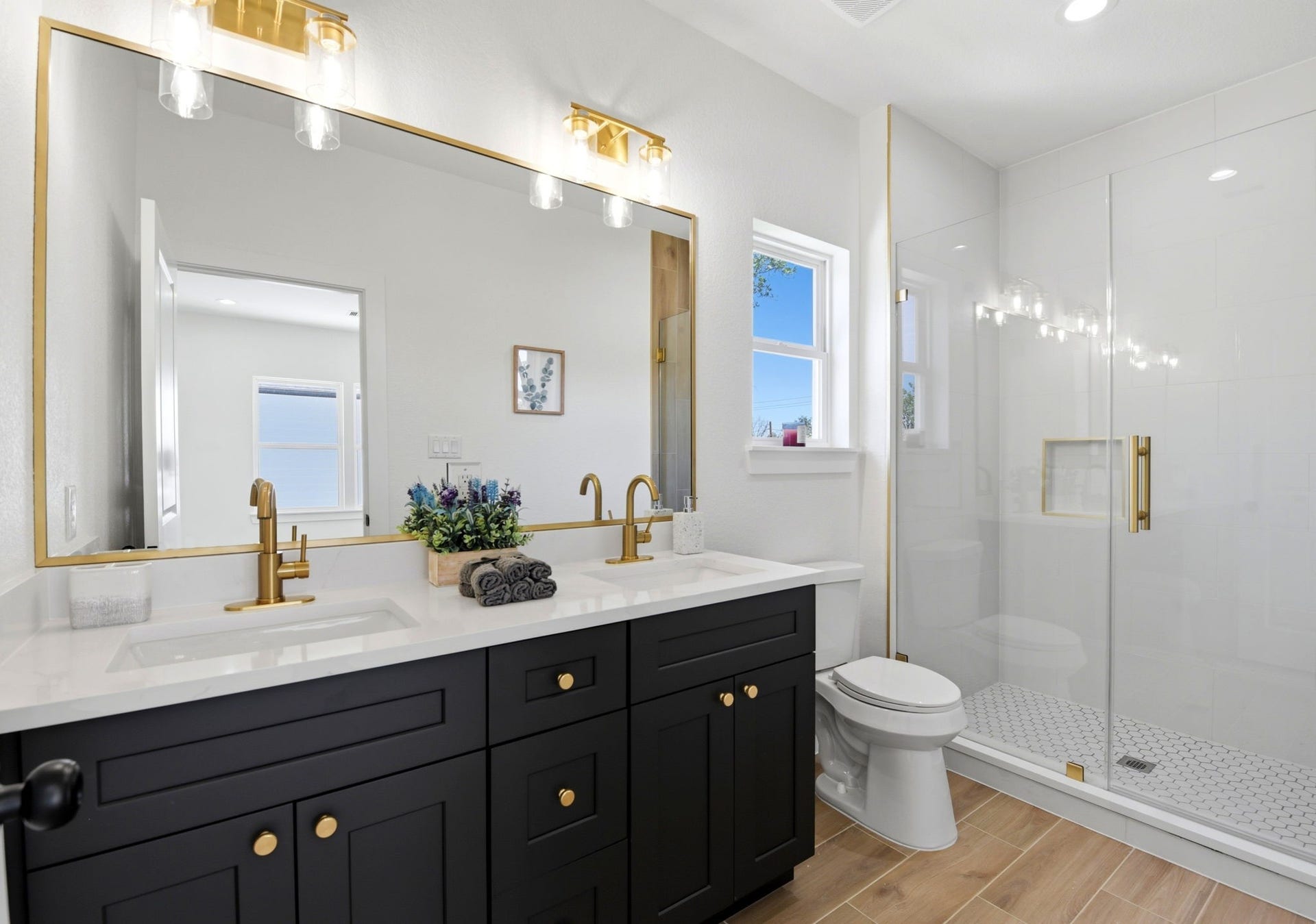 6117 Haight Street, Houston, TX 77028 This modern primary bathroom features a spacious double vanity with sleek black cabinets and gold fixtures. It includes a large mirror, glass-enclosed shower, and bright lighting. The wood-like tile flooring and white walls add warmth and elegance.