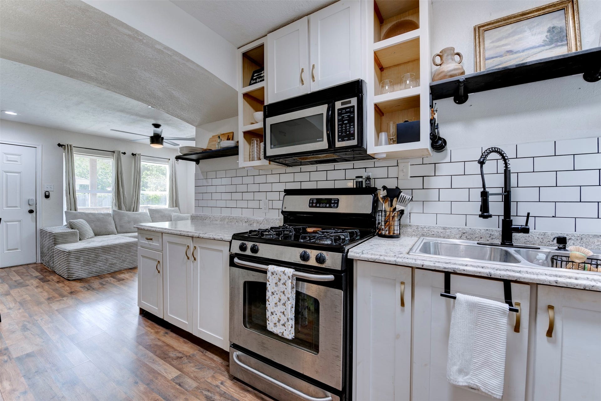 1121 Panama Street, #B, Houston, TX 77009 Kitchen to living room view showcasing GE gas range, stainless over-the-range microwave, matte black pull-down spring faucet, double-basin stainless sink, granite countertops, white subway tile backsplash with dark grout, white shaker cabinetry with brushed gold hardware, open pine cubbies, black pipe bracket floating shelf, dark wood-look LVP flooring, ceiling fan with light kit, and arched opening to living room.