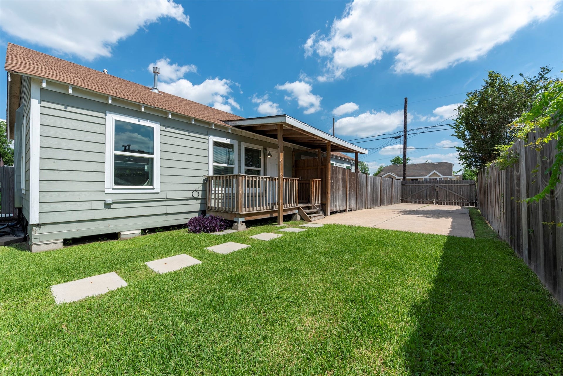 1121 Panama Street, #B, Houston, TX 77009 Side unit view showcasing sage-green lap siding, white-trimmed oversized windows, covered porch with wood post columns, concrete stepping stone path, and gated driveway access.