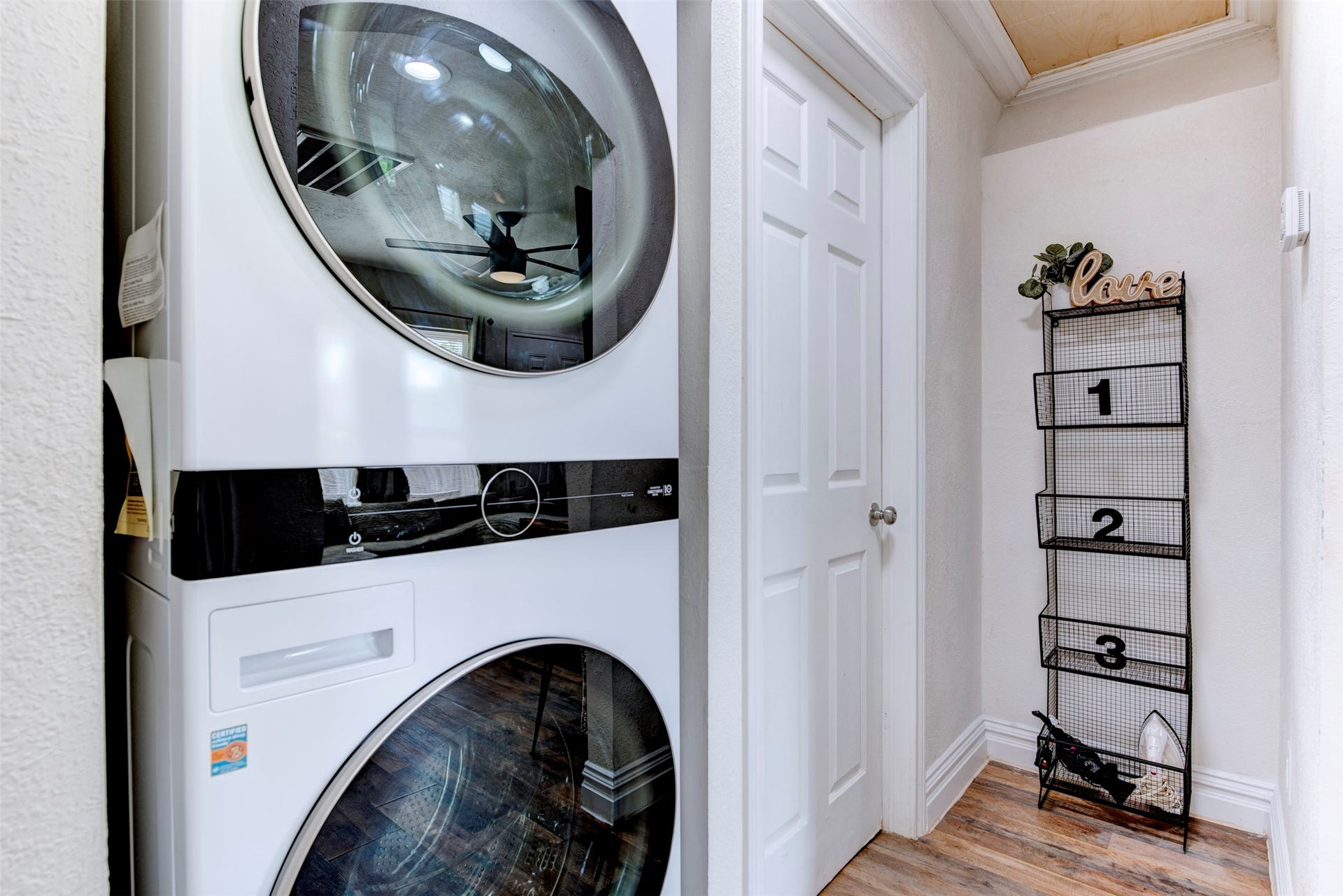 1121 Panama Street, #B, Houston, TX 77009 In-unit hallway laundry closet featuring full-size stacked front-load washer and dryer with black control panel accents, six-panel interior door, wood-look LVP flooring, and wood plank ceiling detail.