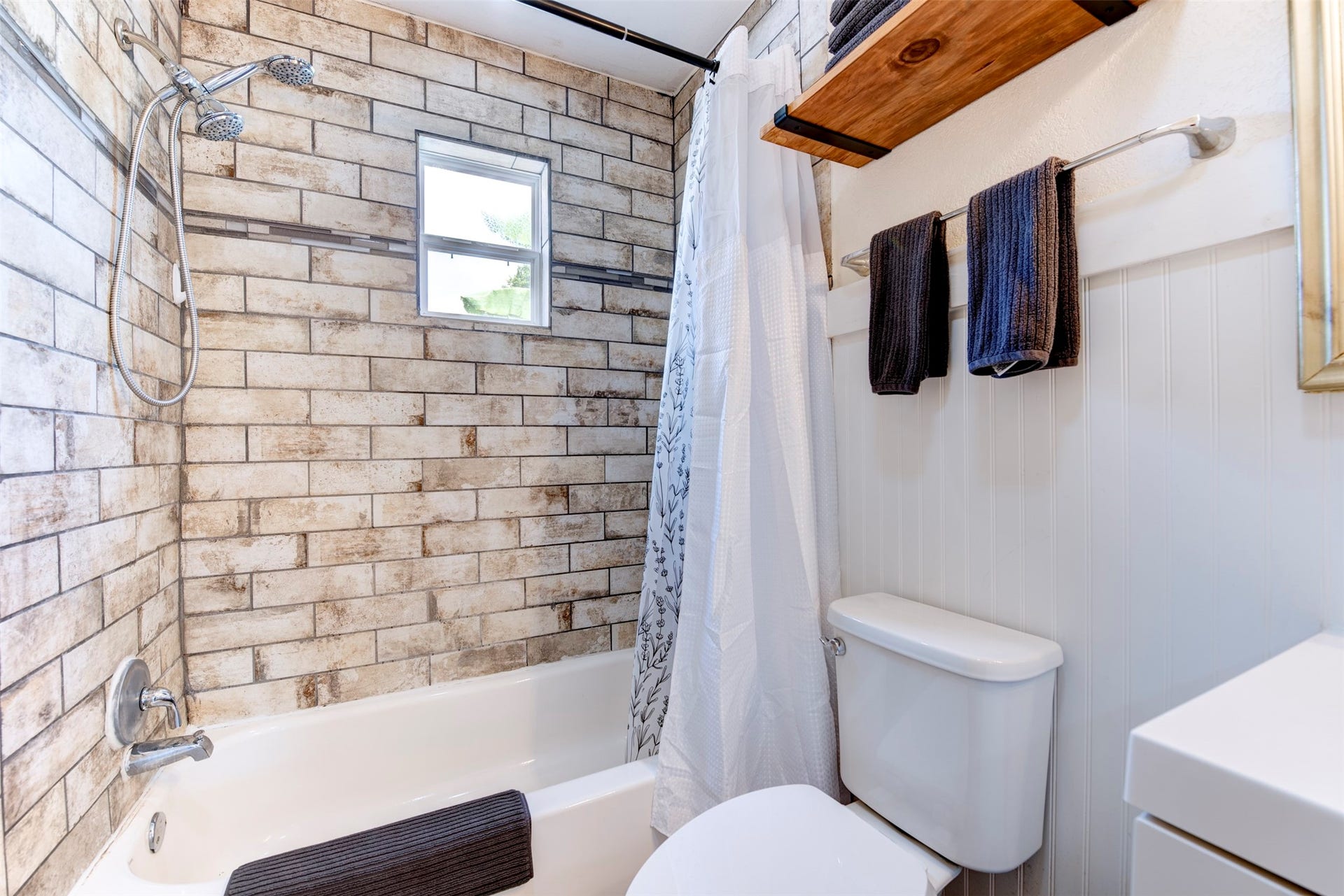1121 Panama Street, #B, Houston, TX 77009 Tub and shower detail featuring floor-to-ceiling rustic brick-style large-format tile, chrome handheld shower head with slide bar, white soaking tub, operable window for natural light and ventilation, natural wood floating shelf with black iron rod, and beadboard wainscoting.