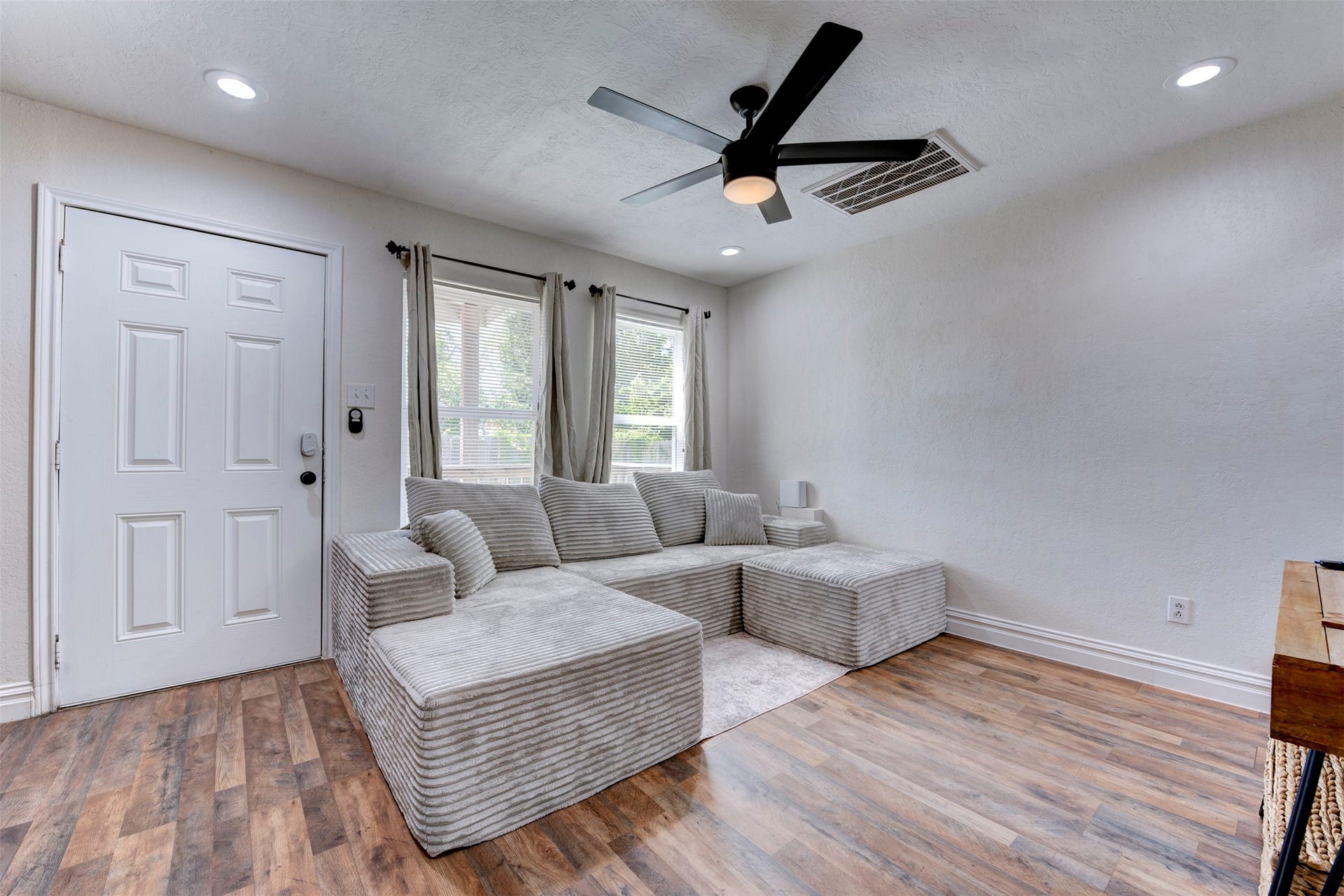 1121 Panama Street, #B, Houston, TX 77009 Living room wide angle showcasing white six-panel entry door with smart lock, oversized window with grommet linen curtains, wood-look LVP flooring, five-blade black ceiling fan with light kit, recessed lighting, and oversized returns with white baseboard trim throughout.
