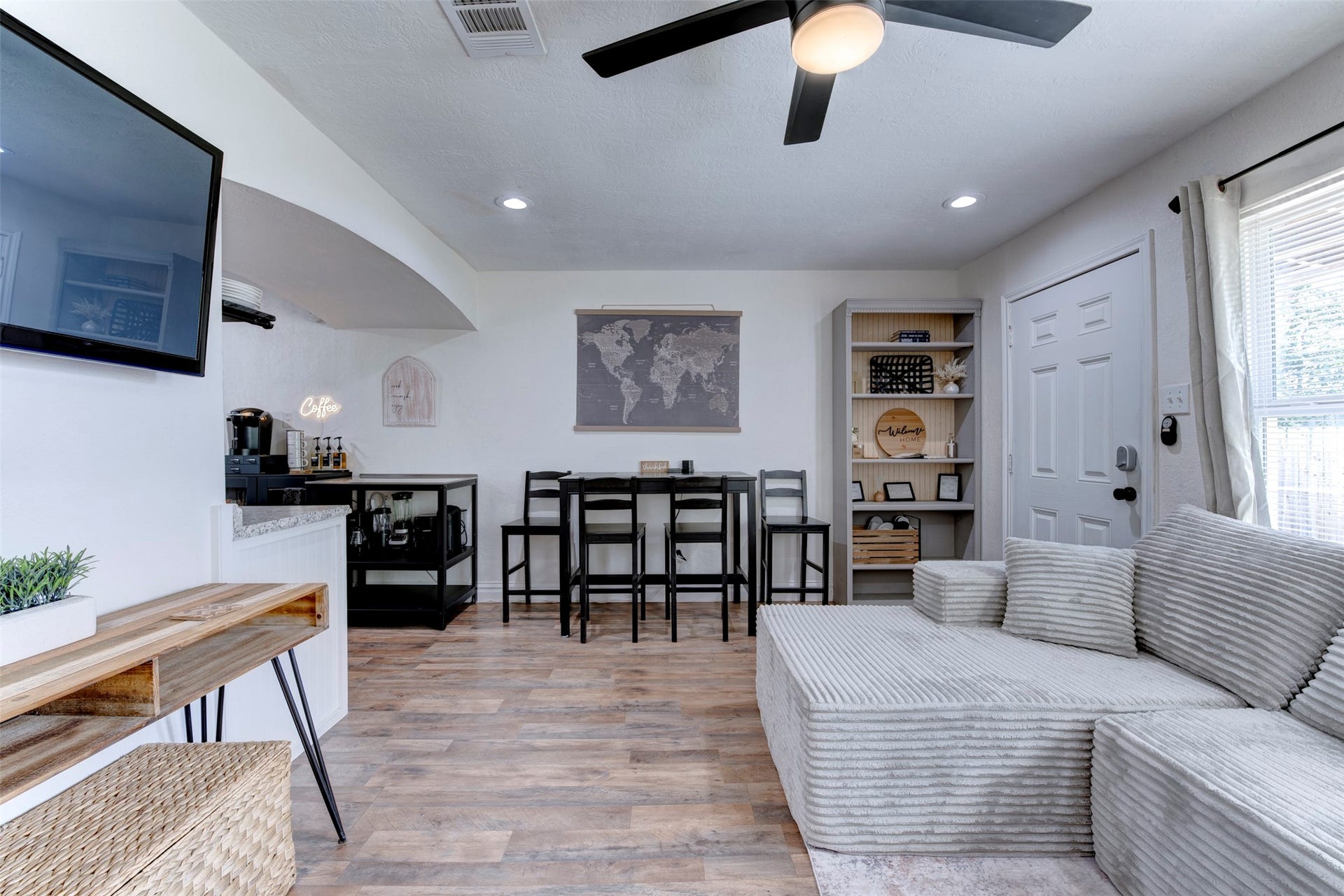 1121 Panama Street, #B, Houston, TX 77009 Full living and dining layout showing wood-look LVP flooring, five-blade black ceiling fan with light kit, recessed lighting, wall-mounted flat screen TV, built-in five-shelf bookcase with beadboard back panel, black bar-height dining set, dedicated coffee station with neon sign, white six-panel entry door with smart lock, and oversized window with natural light.