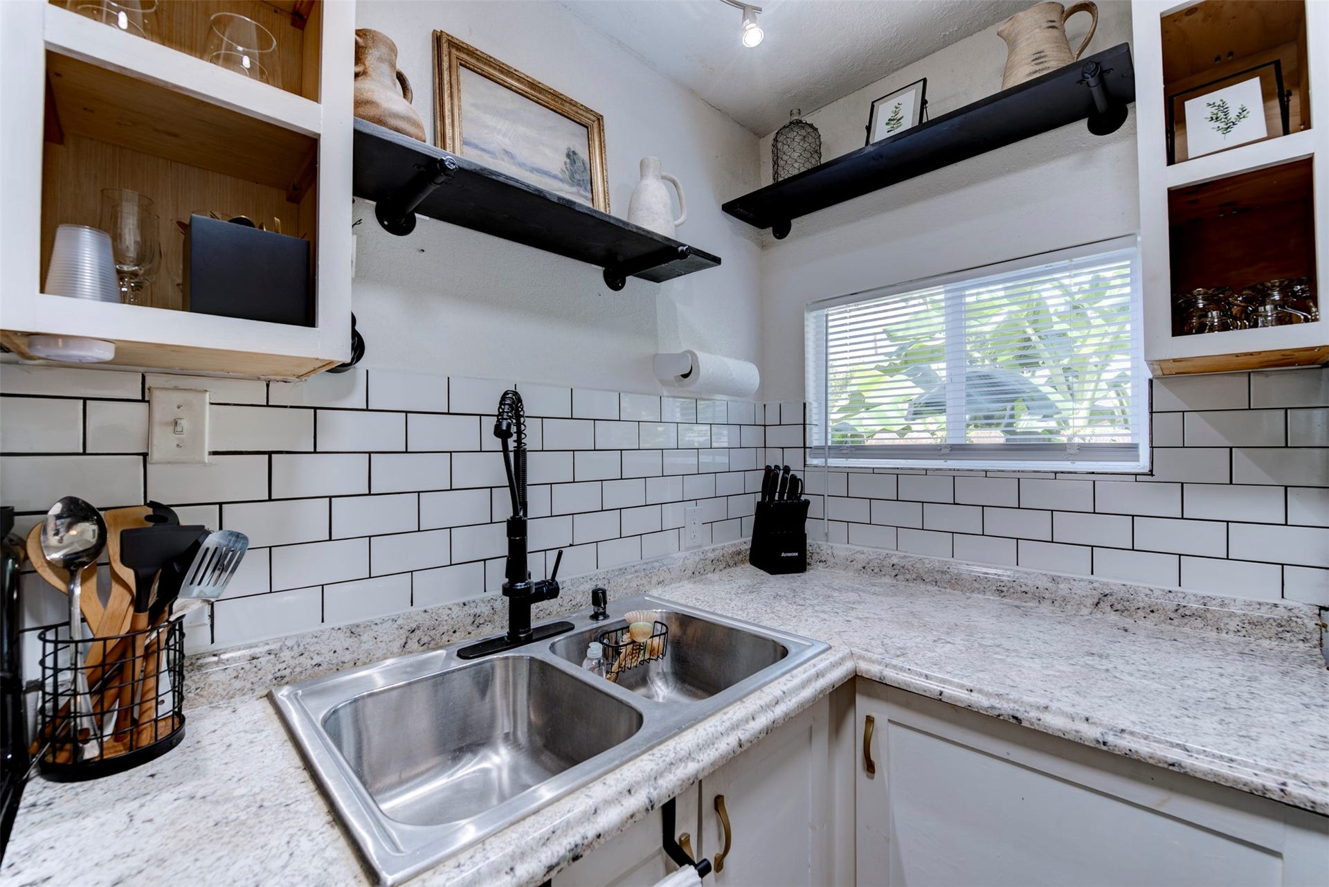 1121 Panama Street, #B, Houston, TX 77009 Kitchen sink area featuring double-basin stainless steel undermount sink, matte black pull-down spring faucet, granite countertops, white subway tile backsplash with dark grout, track lighting, open wood cabinets with natural pine interiors, and dual black pipe bracket floating shelves.