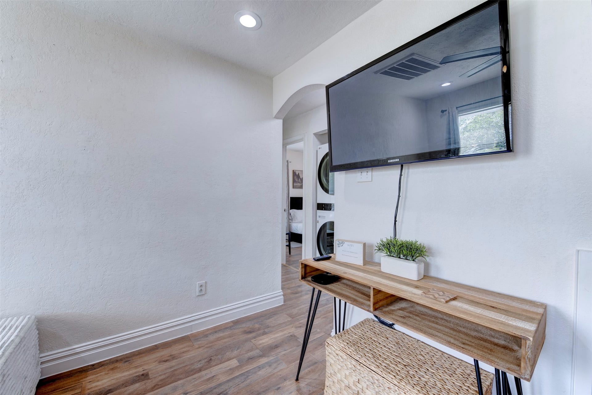 1121 Panama Street, #B, Houston, TX 77009 Living room TV wall detail featuring wall-mounted Samsung flat screen TV, natural wood hairpin leg console table, recessed lighting, arched hallway leading to bedrooms and in-unit stacked laundry, and wood-look LVP flooring throughout.