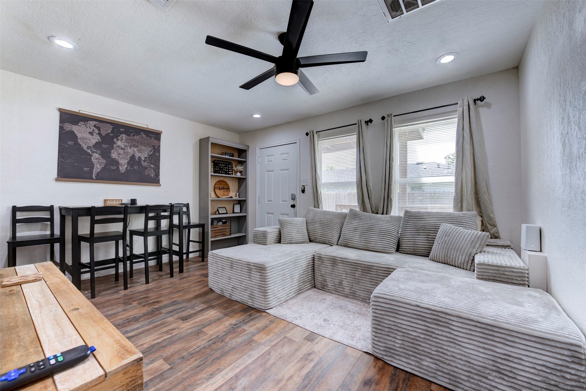 1121 Panama Street, #B, Houston, TX 77009 Living and dining area featuring dark wood-look LVP flooring, oversized black ceiling fan with light kit, recessed lighting, dual oversized windows with grommet linen curtain panels on black rod hardware, five-shelf built-in bookcase with beadboard back panel, six-panel entry door with smart lock, and black bar-height dining set.