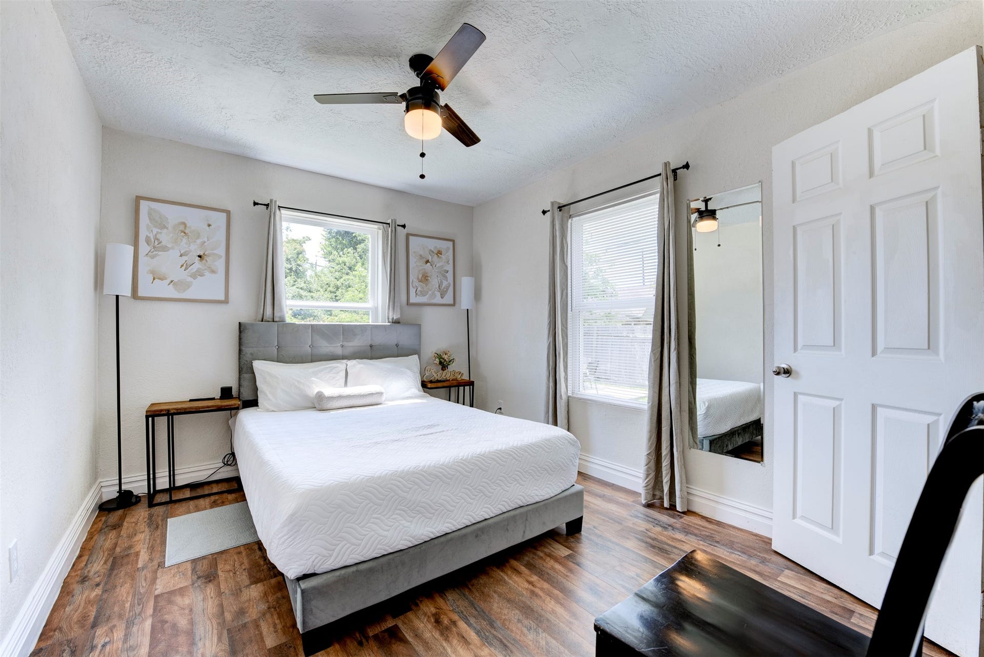 1121 Panama Street, #B, Houston, TX 77009 Primary bedroom with dark wood-look LVP flooring, ceiling fan with light kit, two windows with grommet curtain panels, full-length mirror, and door leading to private bath.