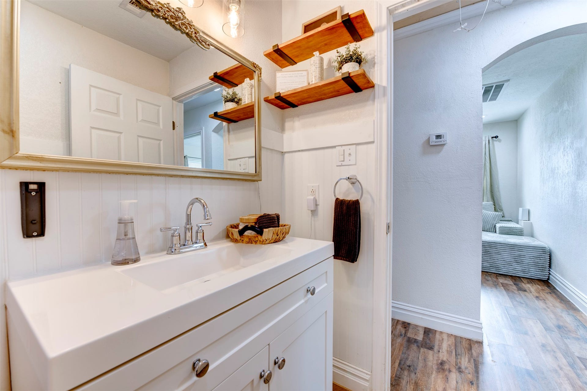 1121 Panama Street, #B, Houston, TX 77009 Bathroom vanity close-up showing white shaker cabinetry with chrome knobs, single-basin white countertop, chrome bridge faucet, ornate silver-framed mirror, dual natural wood floating shelves with black iron brackets, beadboard wainscoting, and open arched hallway leading to bedroom.