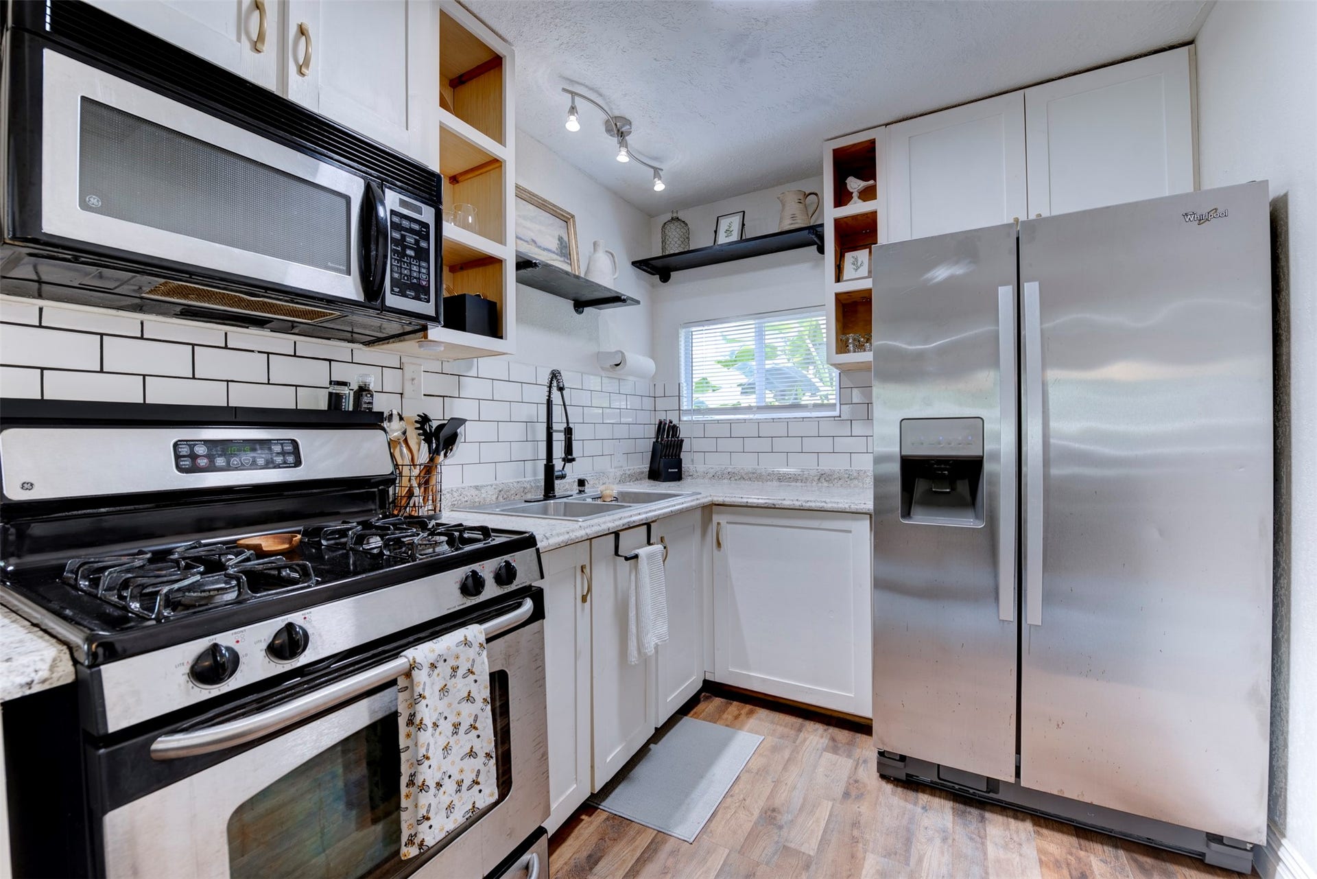 1121 Panama Street, #B, Houston, TX 77009 Full kitchen view featuring GE five-burner gas range with stainless finish, over-the-range stainless microwave, Whirlpool side-by-side refrigerator with ice and water dispenser, white shaker cabinetry with brushed gold hardware, granite countertops, white subway tile backsplash with dark grout, open pine cubby shelving, black pipe bracket floating shelf, track lighting, and operable window.