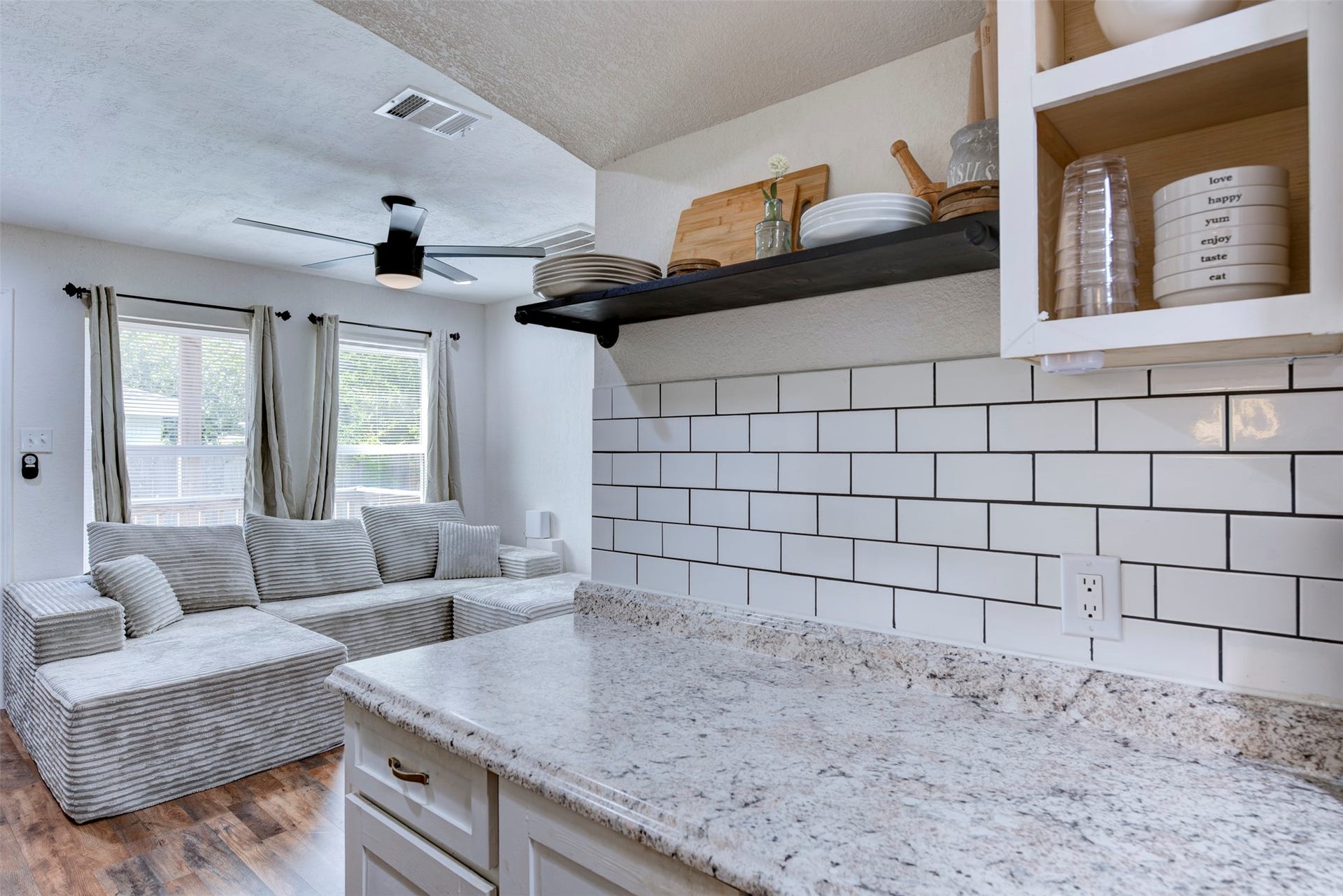 1121 Panama Street, #B, Houston, TX 77009 Kitchen pass-through view showing granite countertops with white and gray veining, white subway tile backsplash with dark grout, black steel open shelving with pipe brackets, white shaker cabinetry with brushed gold hardware, ceiling fan with light kit, and oversized window with grommet curtain panels.