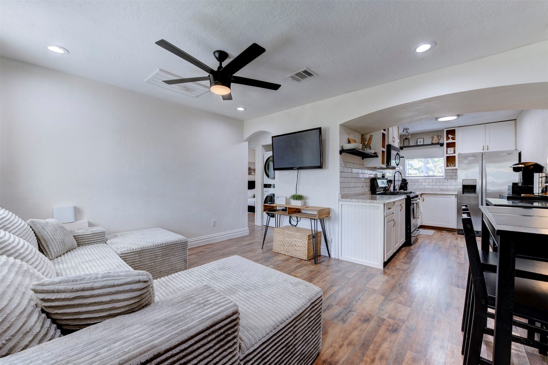 1121 Panama Street, #B, Houston, TX 77009 Open concept living area with wood-look LVP flooring, five-blade black ceiling fan with light kit, recessed lighting, wall-mounted flat screen TV, wood hairpin leg console table, arched opening to hallway and bedrooms, and direct sightlines into updated kitchen with granite countertops, gas range, and stainless refrigerator.