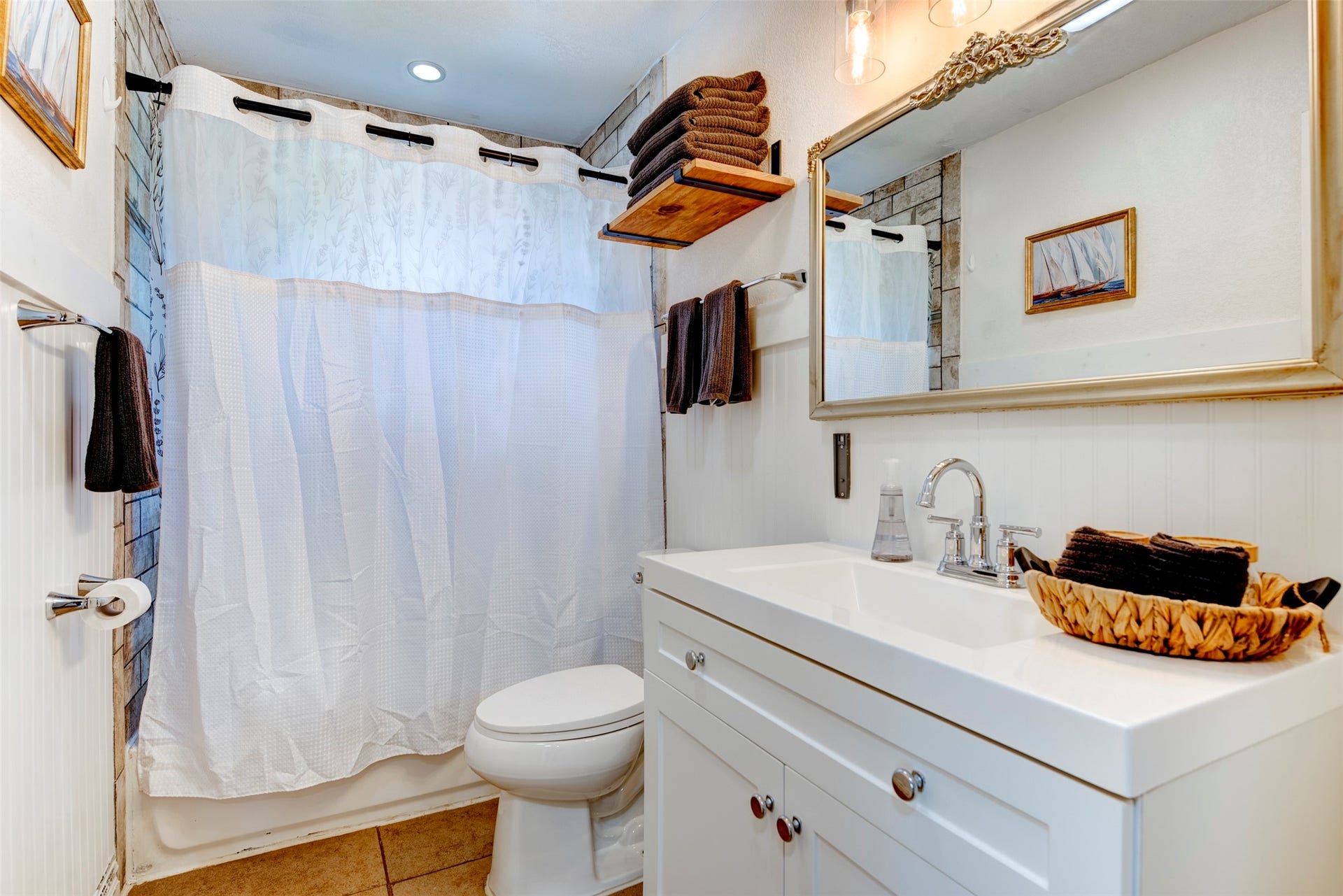 1121 Panama Street, #B, Houston, TX 77009 Full bathroom featuring white shaker vanity with chrome two-handle faucet, ornate silver-framed mirror, Edison bulb pendant lighting, open natural wood floating shelf, beadboard wainscoting, recessed ceiling light, and tiled shower surround.