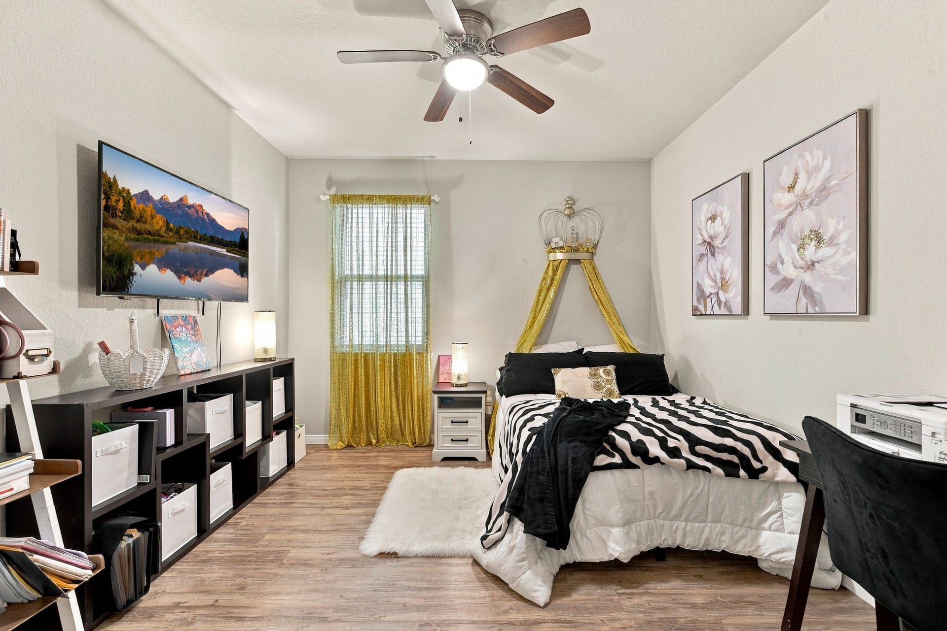 11371 Dawn Beach Lane, Conroe, TX 77304 Cozy bedroom with stylish decor features a comfortable bed with a zebra print throw, large wall-mounted TV, and ample storage with cubbies. Natural light streams through a window with vibrant yellow curtains, complementing the tasteful floral artwork.