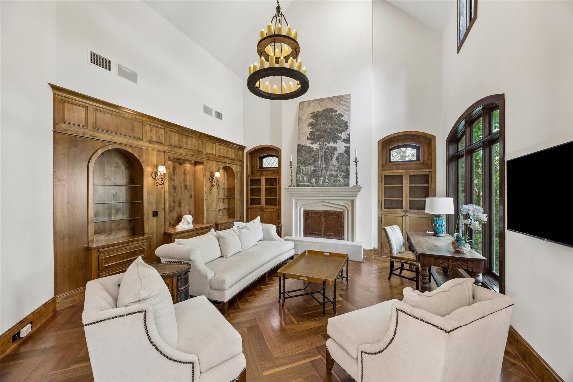 34 Liberty Bell Circle, Bunker Hill Village, TX 77024 Impressive living room featuring soaring ceilings, rich wood-paneled walls with arched built-ins, a carved fireplace mantel, herringbone wood floors, and tall arched windows providing abundant natural light.
