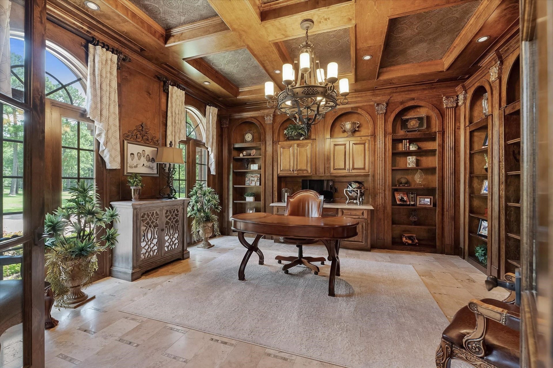 28322 Meadow Forest, Magnolia, TX 77355 This study/library is fully paneled with mahogany wood and features extensive millwork on all walls.
It includes a built-in desk with custom cabinetry that offers lower closed storage and open shelving.
The room boasts a custom coffered tray ceiling with cove lighting, recessed lighting, a designer chandelier, and custom crown molding. Windows provide views of the front lawn with northern exposure, adorned with custom window coverings.
Access to the foyer is through French doors with a valence.