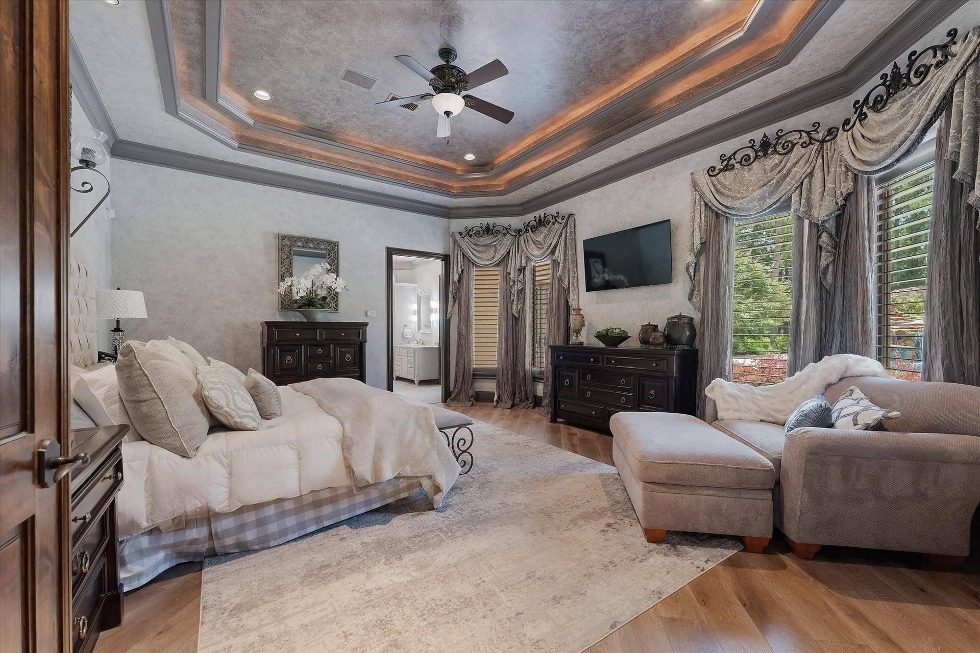 28322 Meadow Forest, Magnolia, TX 77355 Located off the formal living room, the primary bedroom features a dual Adler wood door entry, recessed surround sound speakers, and a custom tray ceiling with cove lighting. Large windows offer views of the backyard with southern and eastern exposures. The room is adorned with custom designer window coverings, recessed LED lighting, custom wood shutters, hardwood floors, designer neutral wall paint, and custom crown molding, creating an elegant and inviting space.