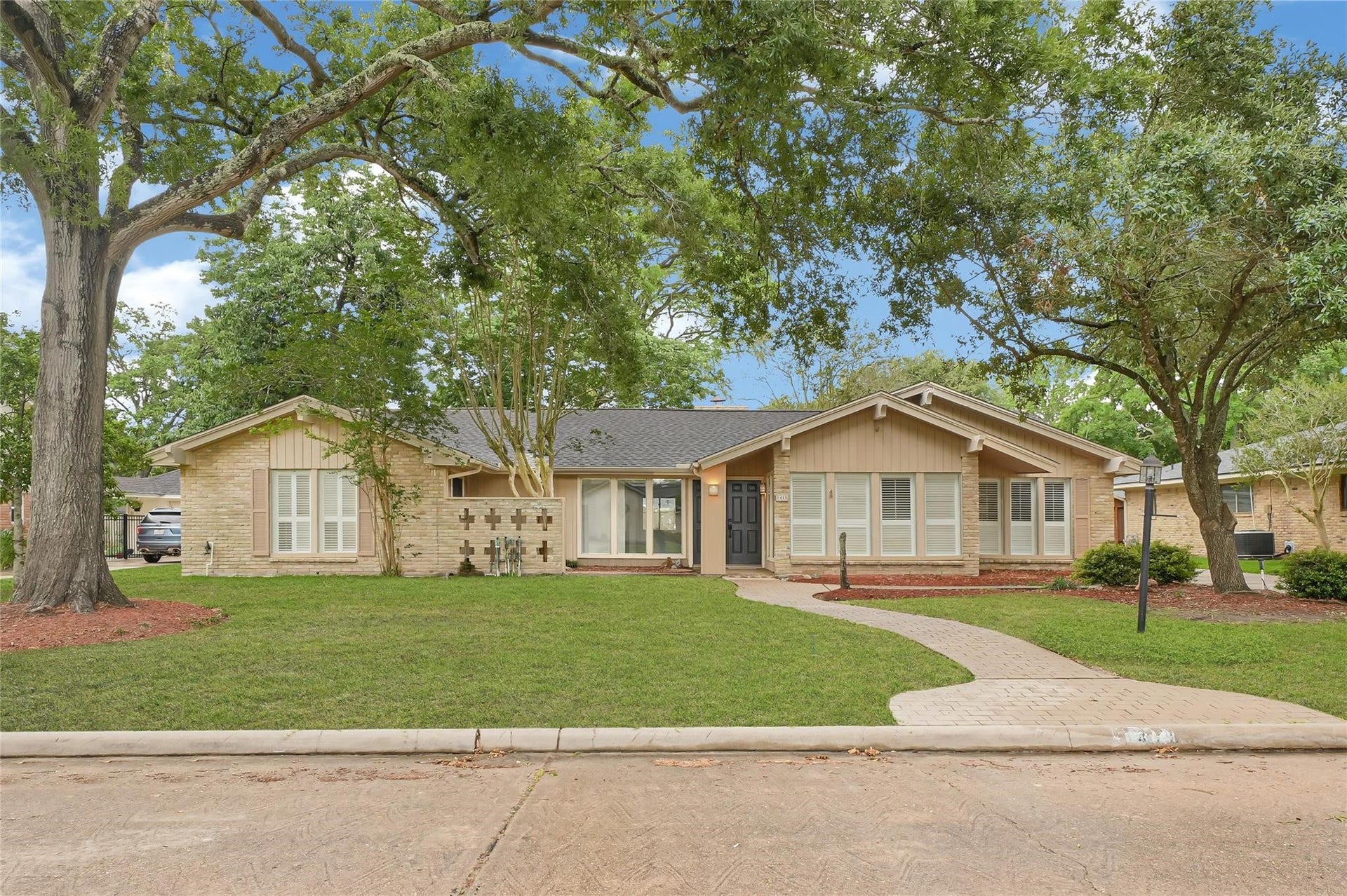 Charming single-story home with a brick exterior, featuring a well-maintained lawn and mature trees. The inviting entrance includes a paved walkway leading to a classic black front door. Large windows provide ample natural light.