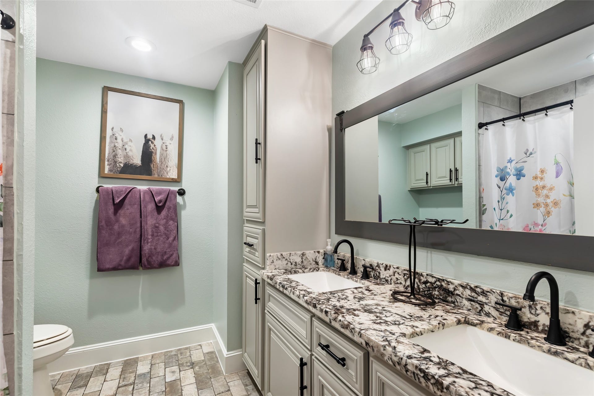 25107 Goodson Road, Splendora, TX 77372 Charming bathroom with a dual-sink granite vanity, elegant black fixtures, and stylish lighting. The soft color palette complements the floral shower curtain and decorative wall art, creating a cozy and inviting atmosphere.