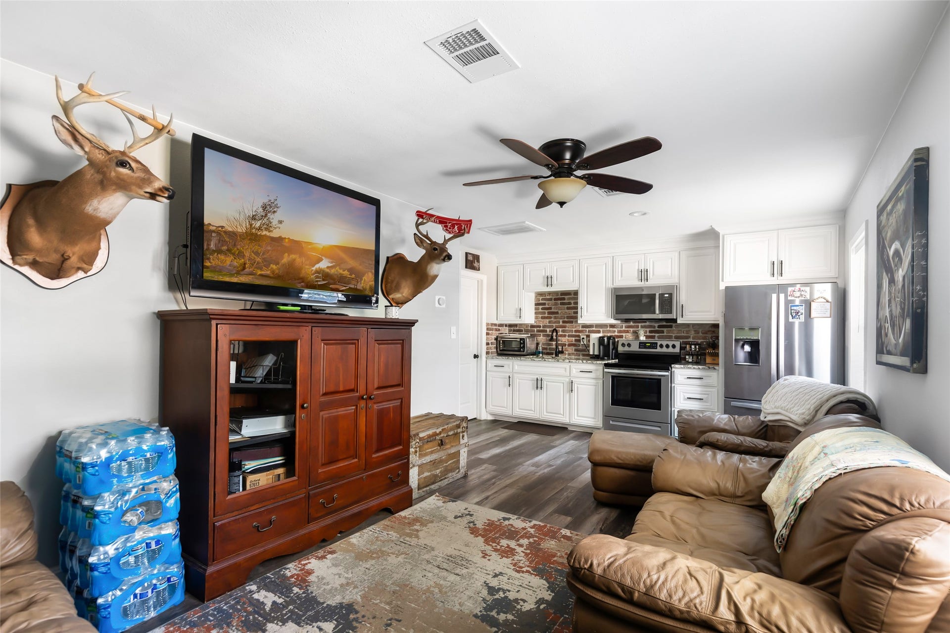 25107 Goodson Road, Splendora, TX 77372 Cozy living area with an open-plan design that seamlessly flows into a modern kitchen featuring stainless steel appliances, white cabinetry, and a stylish brick backsplash. Plush leather seating and a ceiling fan provide comfort, while decorative accents add character.