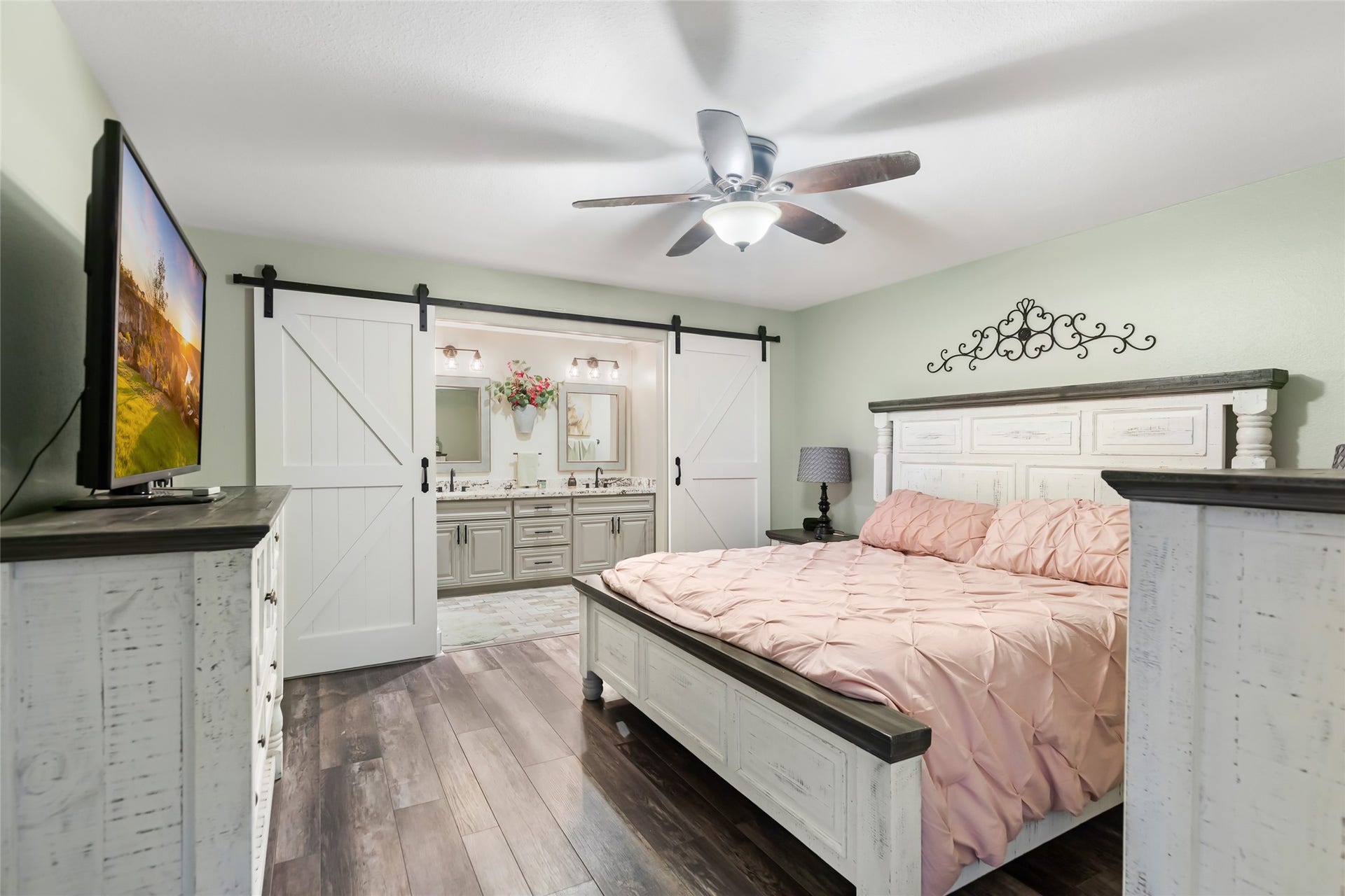25107 Goodson Road, Splendora, TX 77372 Charming bedroom with rustic barn doors, elegant wood flooring, and a ceiling fan, leading to a stylish ensuite with dual sinks.