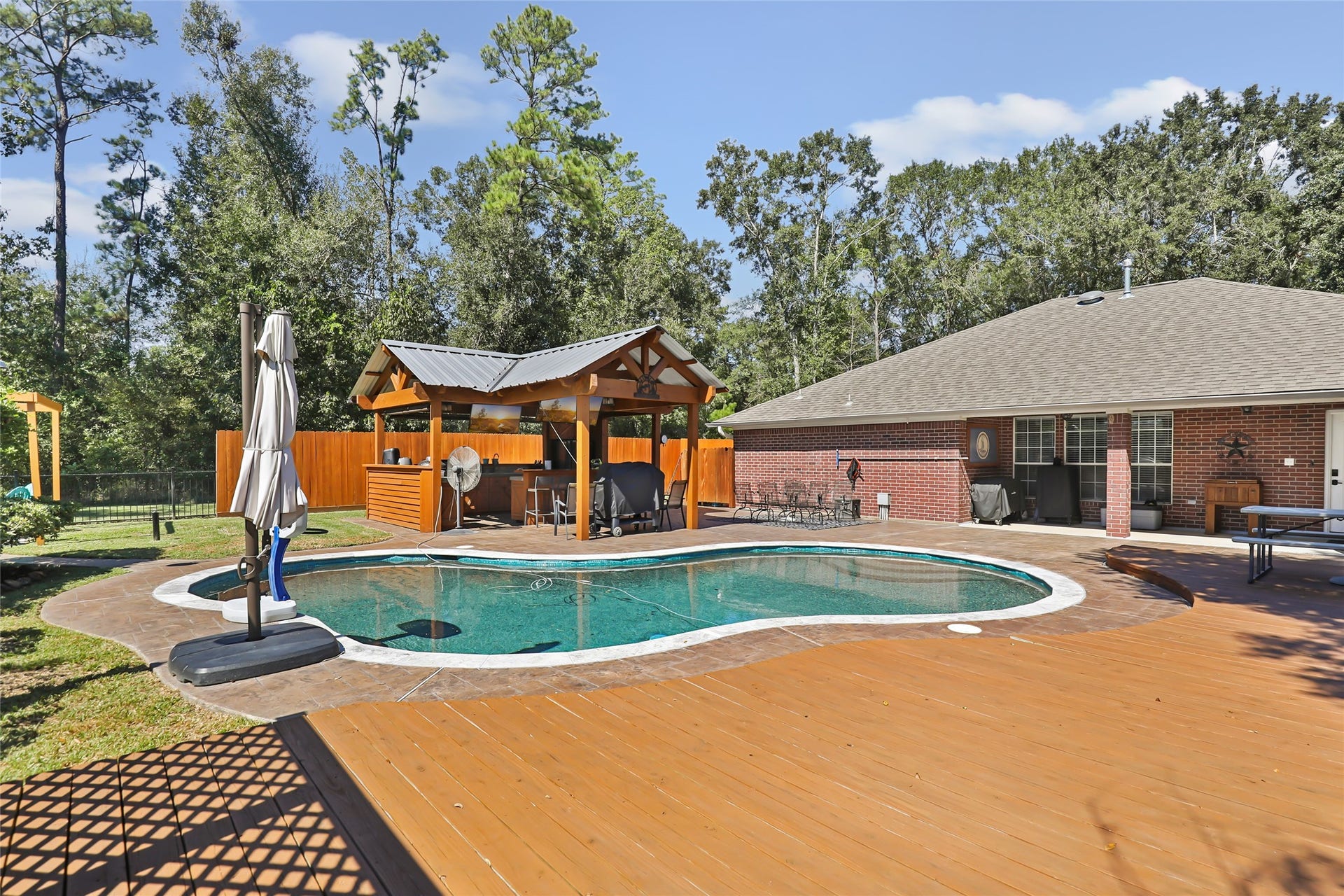 25107 Goodson Road, Splendora, TX 77372 Charming backyard oasis featuring a sparkling pool and a spacious wooden deck, complete with a covered outdoor kitchen and surrounded by lush greenery for privacy and relaxation.
