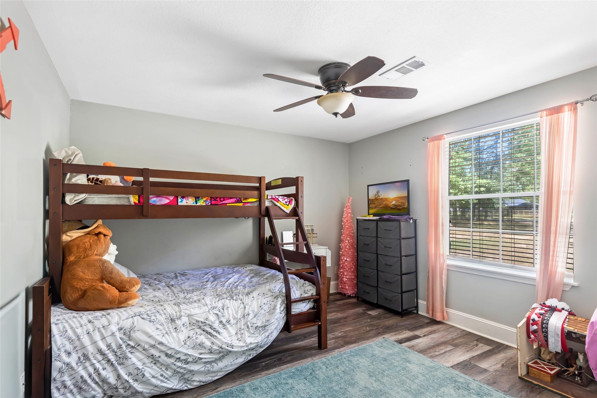25107 Goodson Road, Splendora, TX 77372 Charming bedroom with a cozy bunk bed and a large window allowing abundant natural light. The room features a ceiling fan, soft gray walls, and stylish wood flooring, creating a welcoming atmosphere.