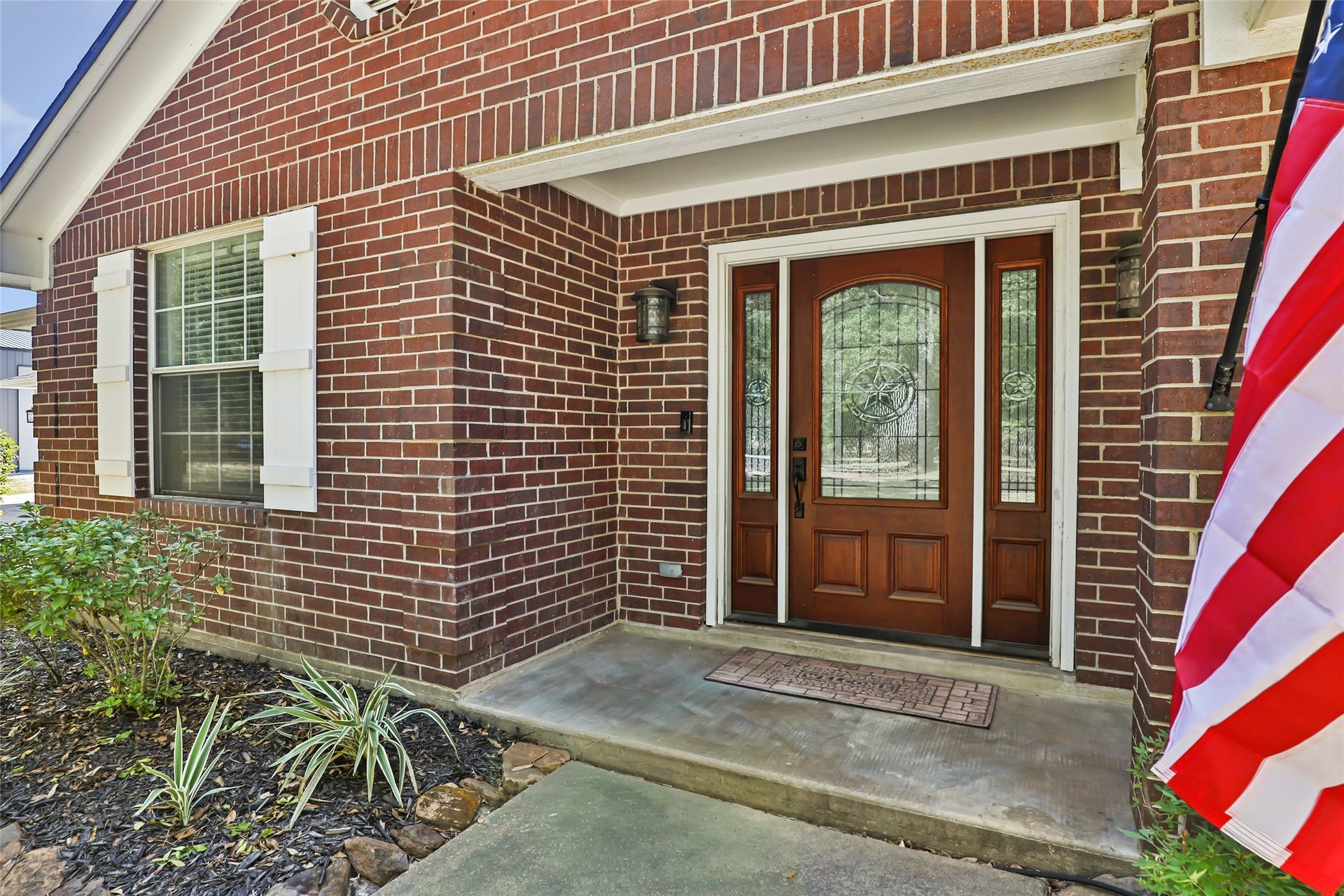 25107 Goodson Road, Splendora, TX 77372 Charming brick exterior with a welcoming front door featuring decorative glass panels, complemented by tasteful landscaping and an inviting flag display.