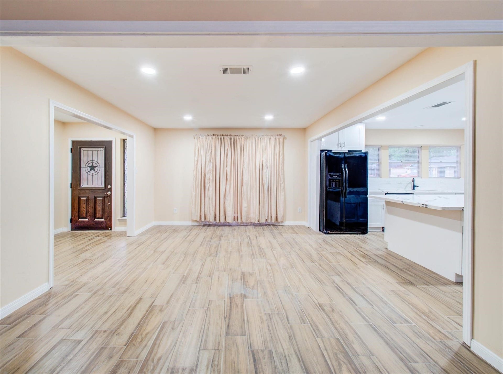 13526 Sherwood Street, Sugar Land, TX 77498 Open-concept living area with light wood flooring, beige walls, and recessed lighting. Features a modern kitchen with black appliances and a spacious layout. A front door with decorative glass and large windows provide natural light.