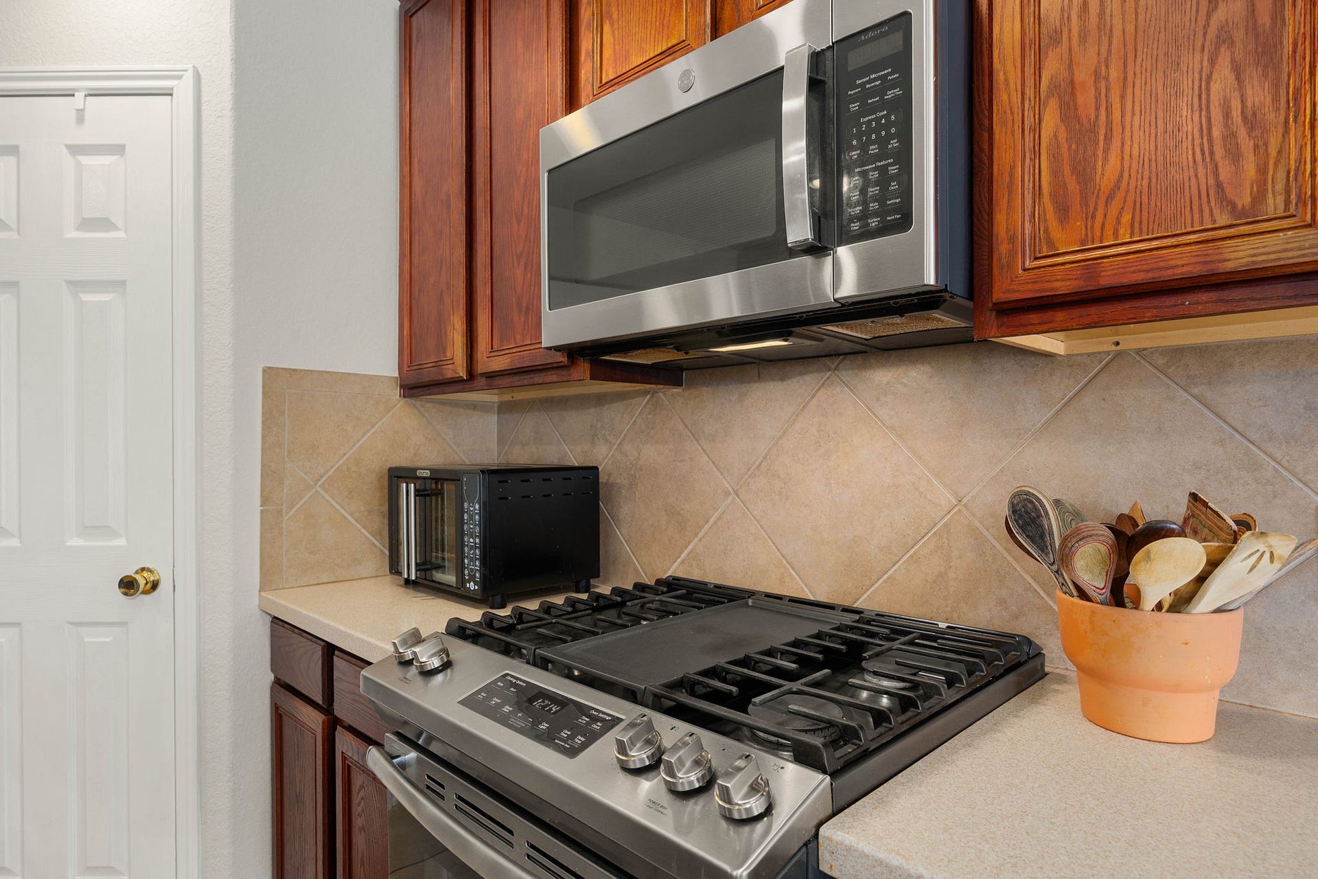 Upgraded cooking zone featuring high-quality stainless steel GE appliances, including a built-in microwave and a modern gas range with a center griddle burner. The space is beautifully tied together by a classic tile backsplash and rich wood cabinetry, offering both functionality and style for the home chef.