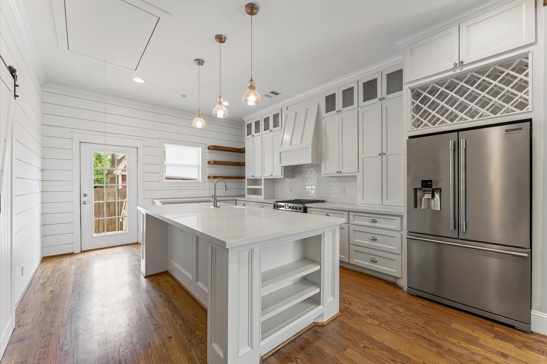 1204 Shearn Street, Houston, TX 77007 This modern kitchen features a spacious layout with white cabinetry, a large center island, stainless steel appliances, and elegant pendant lighting. The room is complemented by hardwood floors and a door leading to the outside, offering ample natural light.