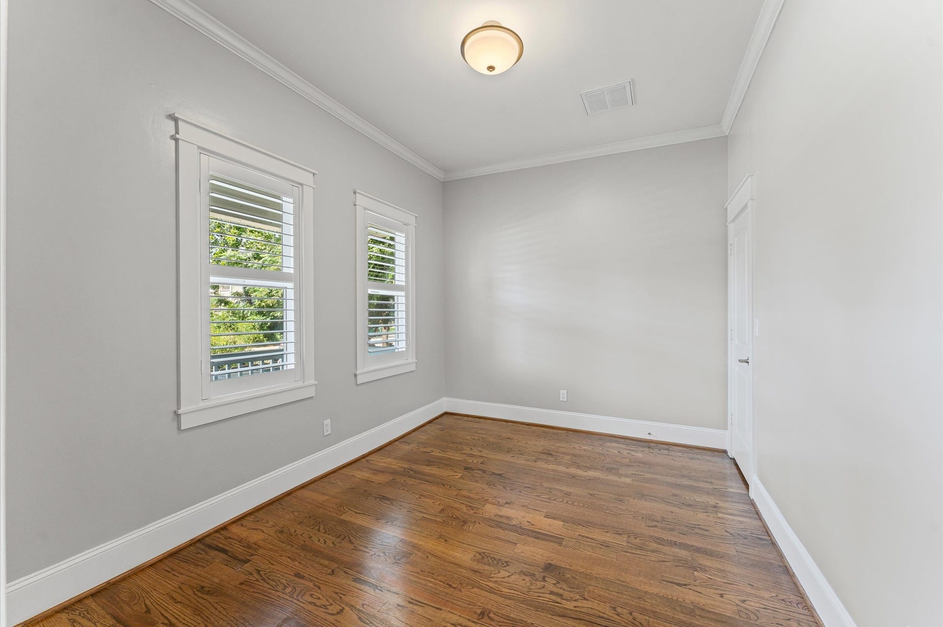 1204 Shearn Street, Houston, TX 77007 Bright room with hardwood floors, light gray walls, and crown molding. Features two windows with shutters, a ceiling light, and a white door. Ideal for a bedroom or office.