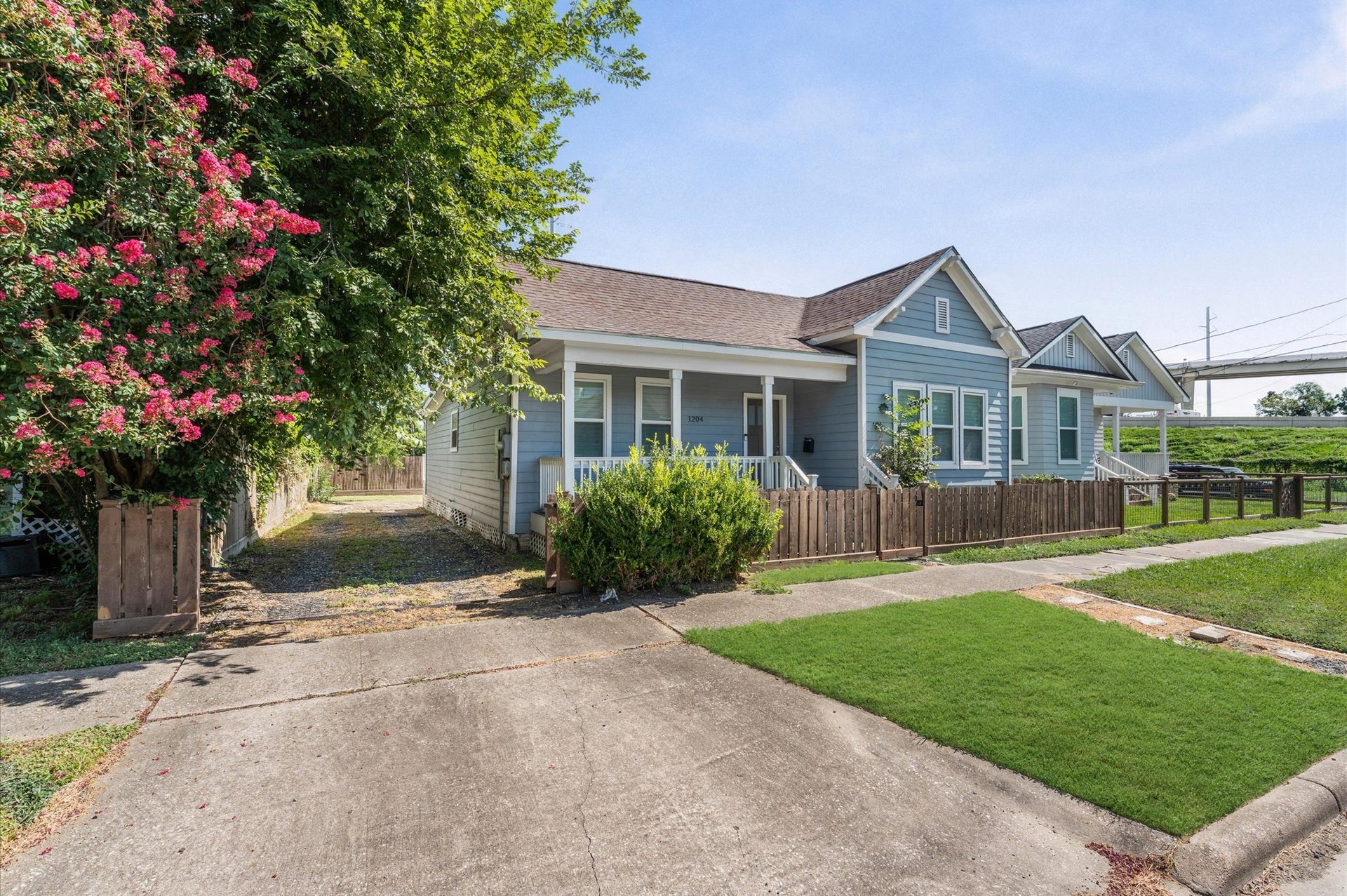 1204 Shearn Street, Houston, TX 77007 Charming blue bungalow with a welcoming front porch, surrounded by a wooden fence and lush greenery. The property features a well-maintained lawn, vibrant flowering trees, and a paved driveway, offering a cozy, inviting atmosphere.
