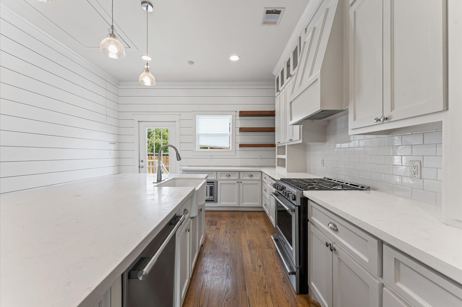 1204 Shearn Street, Houston, TX 77007 This kitchen features a bright, contemporary design with white cabinetry and shiplap walls. It includes modern stainless steel appliances, a farmhouse sink, and a spacious island with pendant lighting. The wooden flooring adds warmth, and there's ample natural light from a window and glass door.