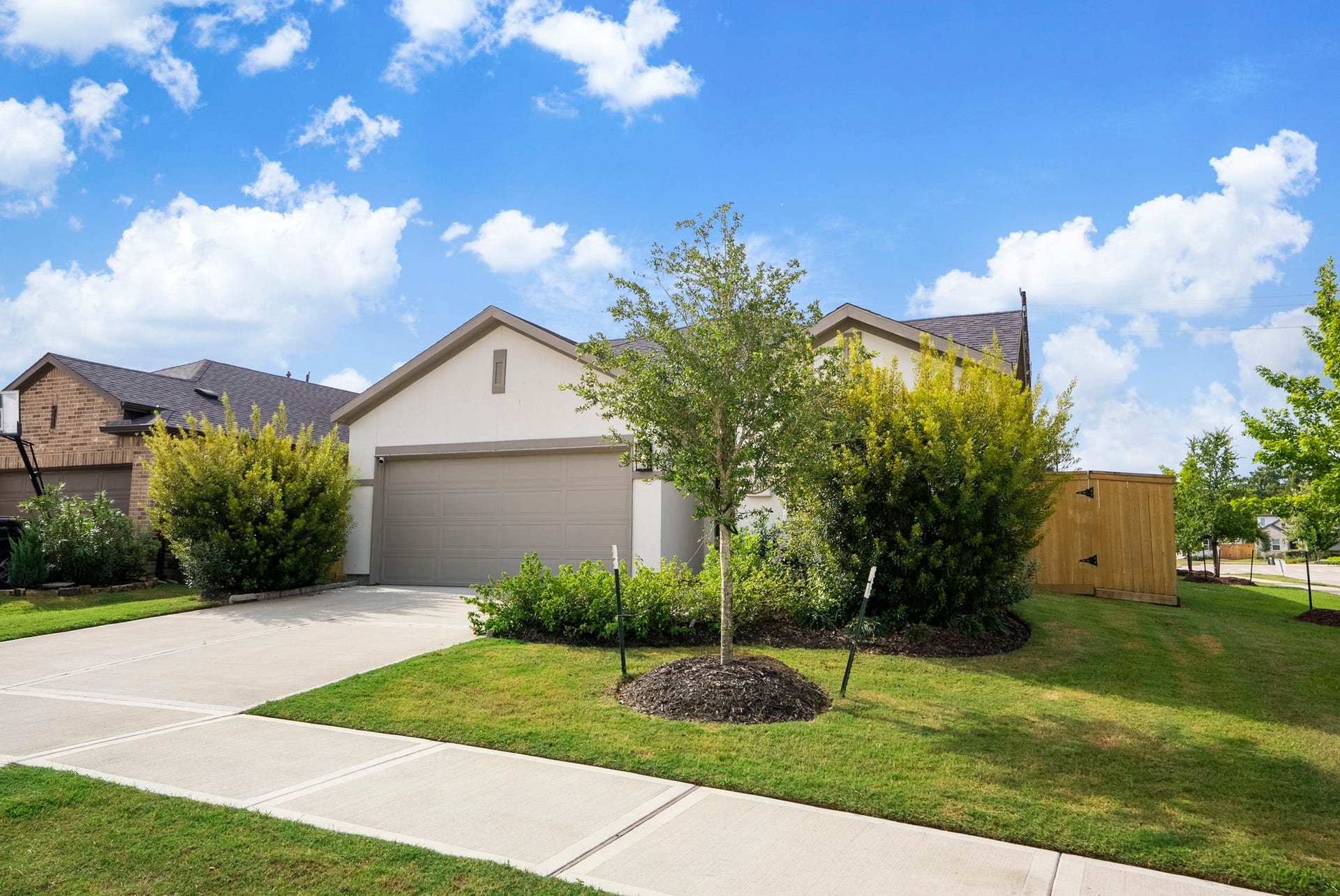 This photo showcases a modern single-story home with a two-car garage, well-maintained front yard, and a concrete driveway. It features lush landscaping with shrubs and a young tree, offering curb appeal in a suburban neighborhood setting.