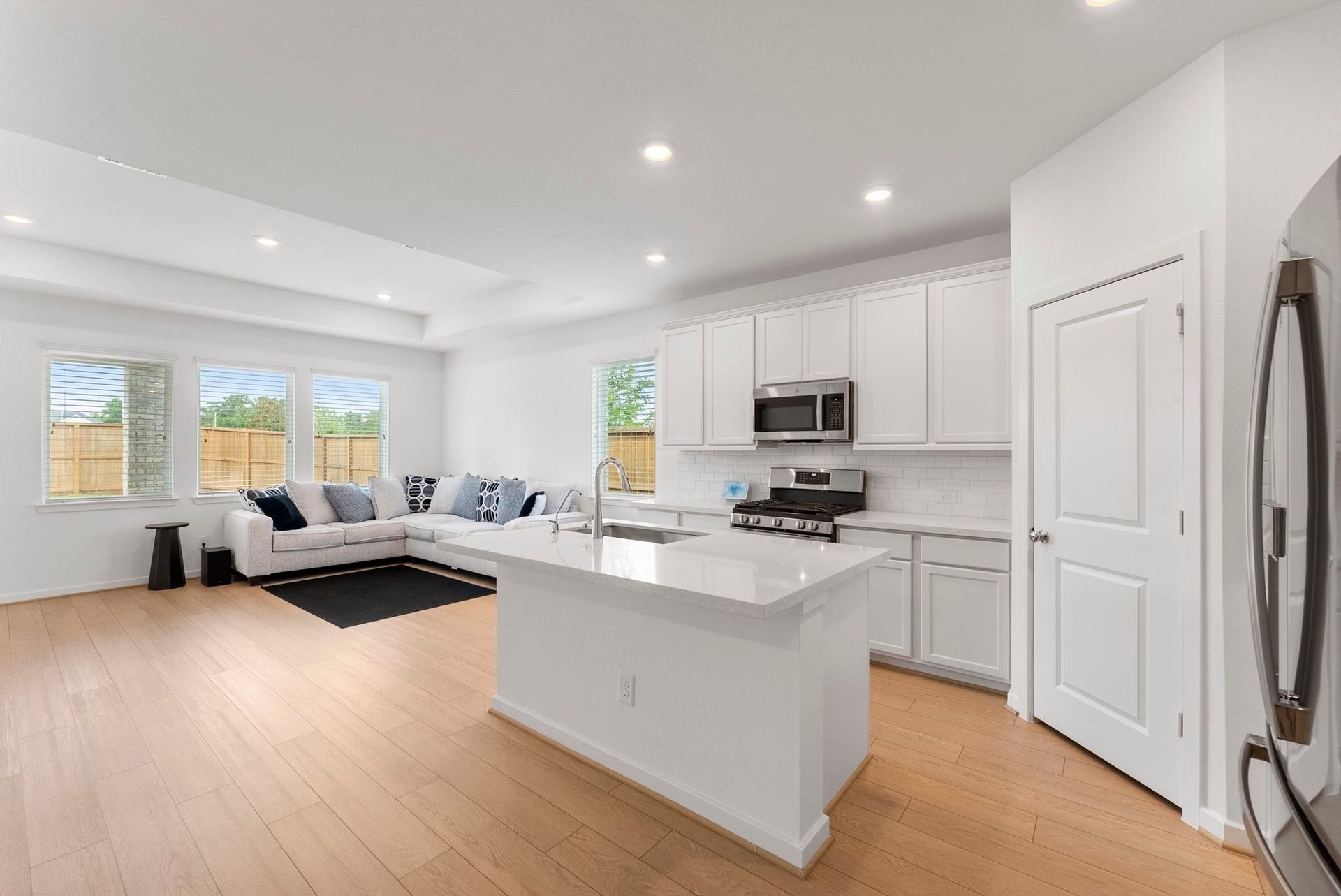 This photo showcases a modern, open-concept kitchen and living area. The kitchen features white cabinetry, stainless steel appliances, and a central island with a sink. The living space includes a cozy sectional sofa and large windows that let in natural light, creating a bright and inviting atmosphere.