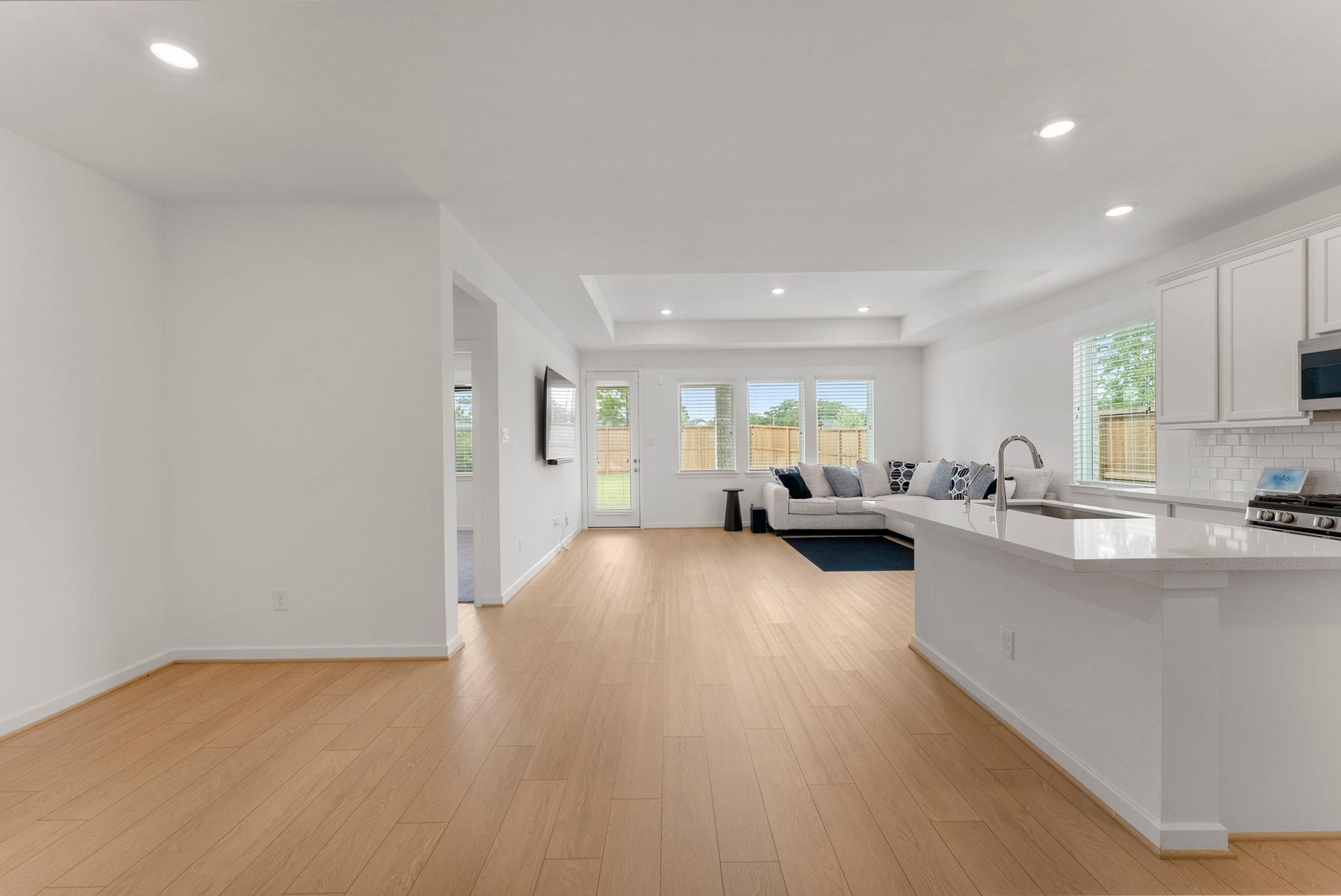 This photo showcases a bright, open-concept living area with modern finishes. The space features light wood flooring, a cozy living room with large windows, and a sleek kitchen with white cabinetry and a breakfast bar.