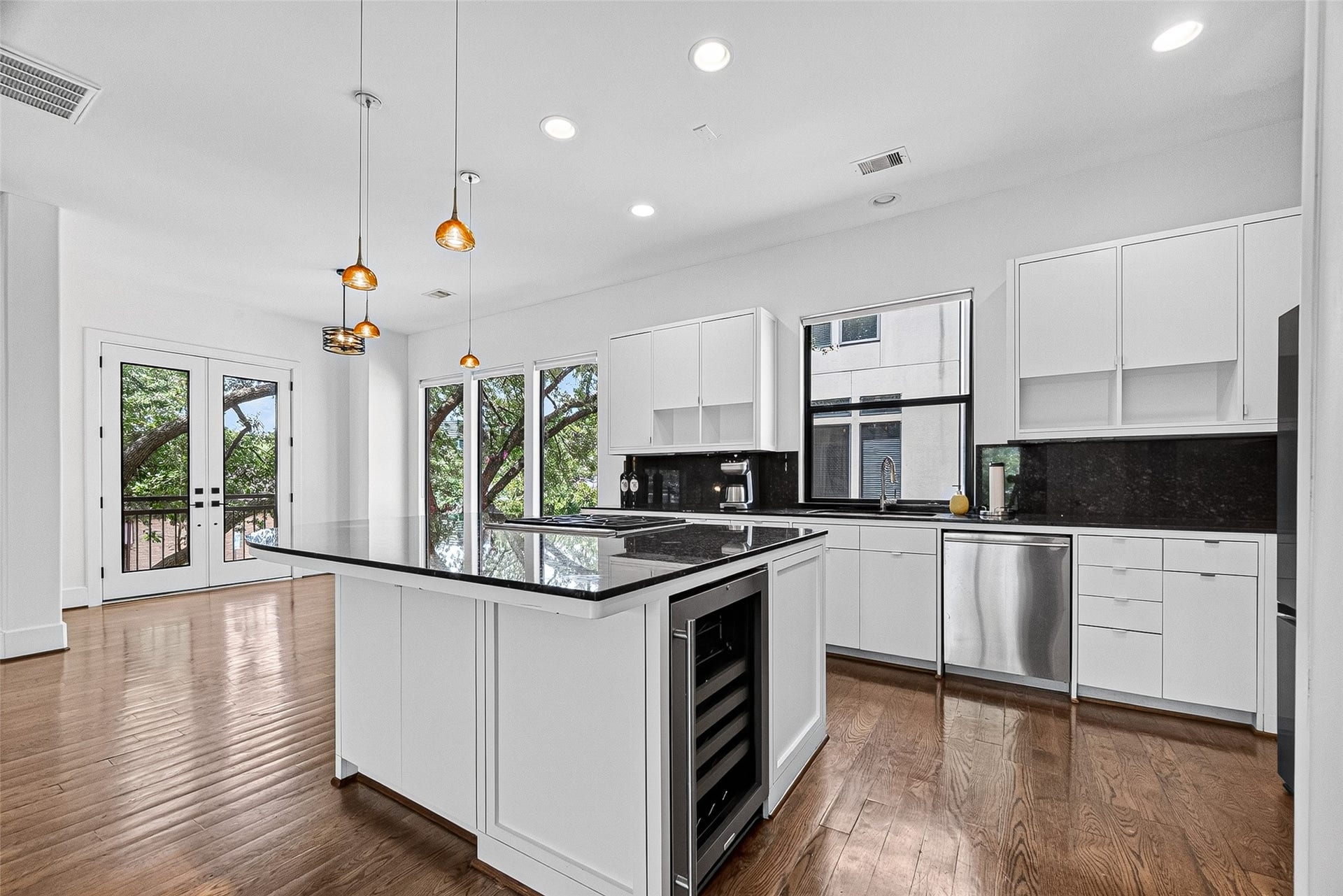 1110 Autrey Street, #C, Houston, TX 77006 Elegant kitchen featuring sleek white cabinetry, a spacious island with a cooktop and wine cooler, and stunning hardwood floors. Abundant natural light streams in through large windows and glass doors, offering a view of lush greenery.