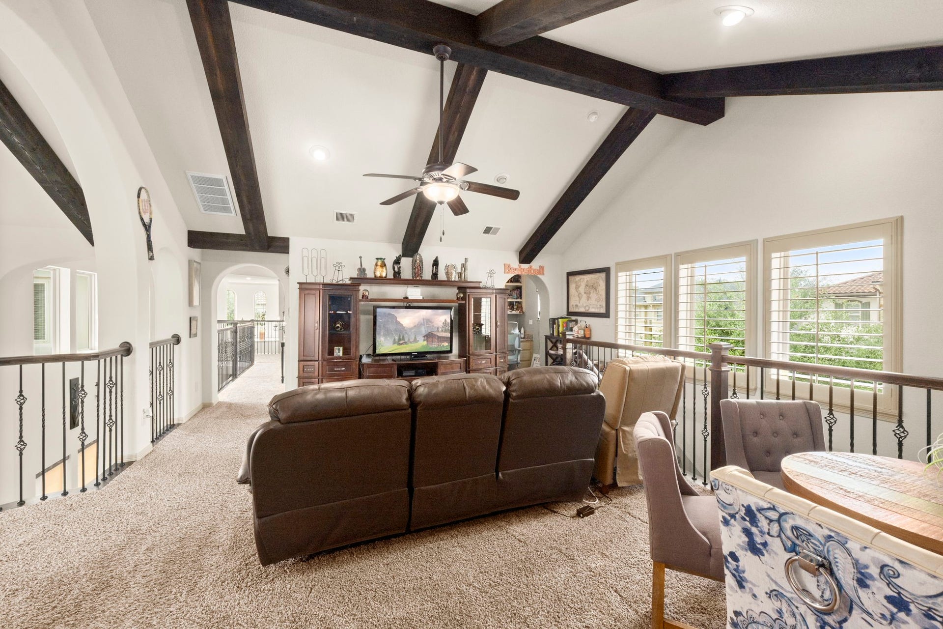 5103 Jackson Ponds Court, Sugar Land, TX 77479 This spacious, carpeted loft features high ceilings with exposed wooden beams, a ceiling fan, and ample natural light from large windows. It includes a cozy seating area with a TV, a railing overlooking the floor below, and a small dining or game table, creating a versatile living space.