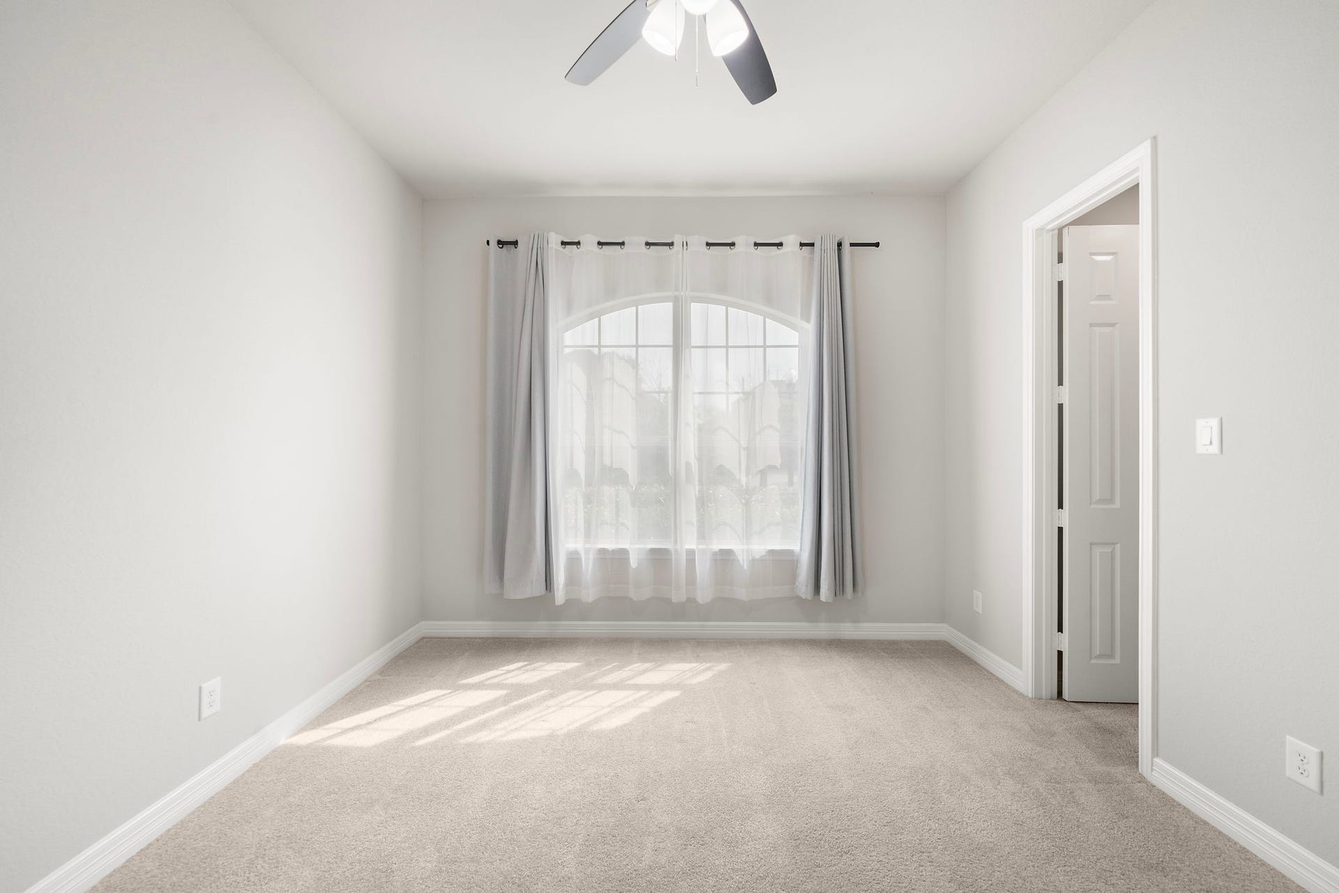 A second view of the bedroom highlights its impressive proportions, soft carpeting, neutral color palette, and beautiful arched window. Natural light enhances the bright atmosphere, while the ceiling fan and convenient adjoining doorway add everyday comfort and functionality to this peaceful private space.