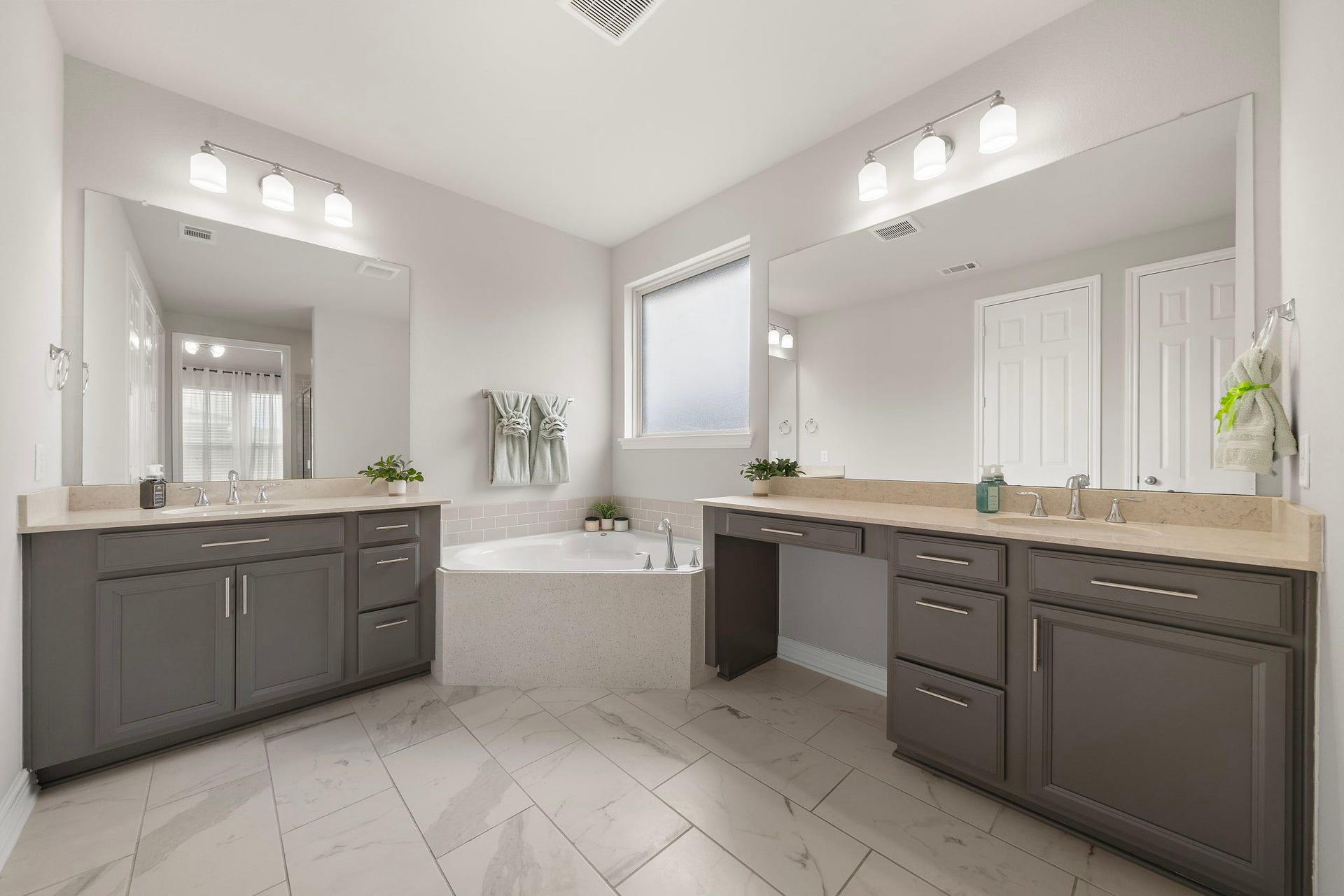 Designed for comfort and convenience, the primary bathroom features dual vanity areas with generous cabinetry, expansive mirrors, and ample counter space. A soaking tub positioned beneath a frosted window adds a relaxing focal point, while the neutral tile flooring and coordinated finishes create a polished, cohesive look.