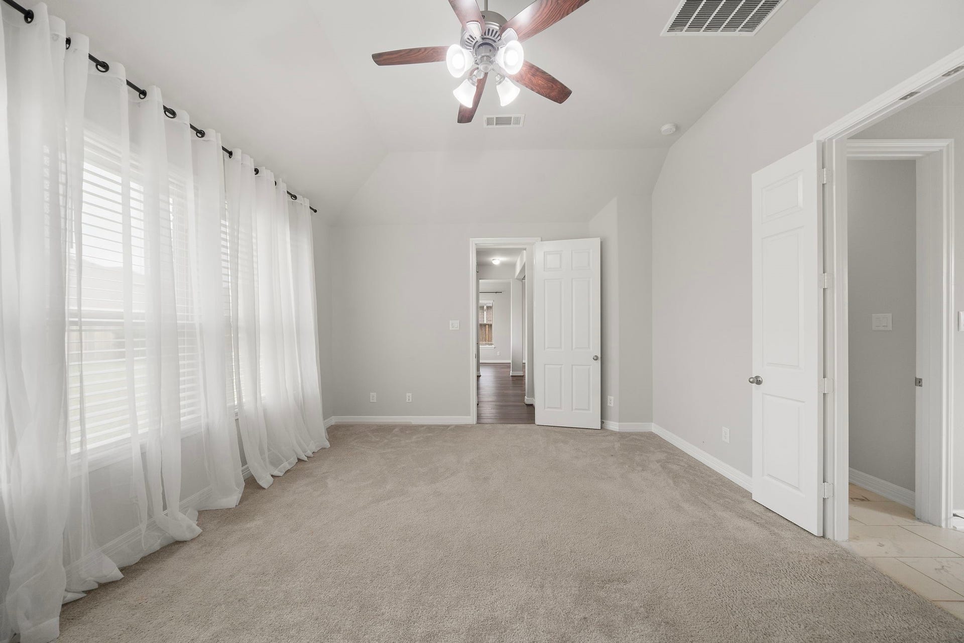 This wide bedroom view showcases the generous proportions of the primary suite along with its soft neutral palette, plush carpeting, and expansive windows. The ceiling fan provides added comfort, while convenient access to the adjoining bath and hallway creates a functional layout designed for privacy and everyday ease.