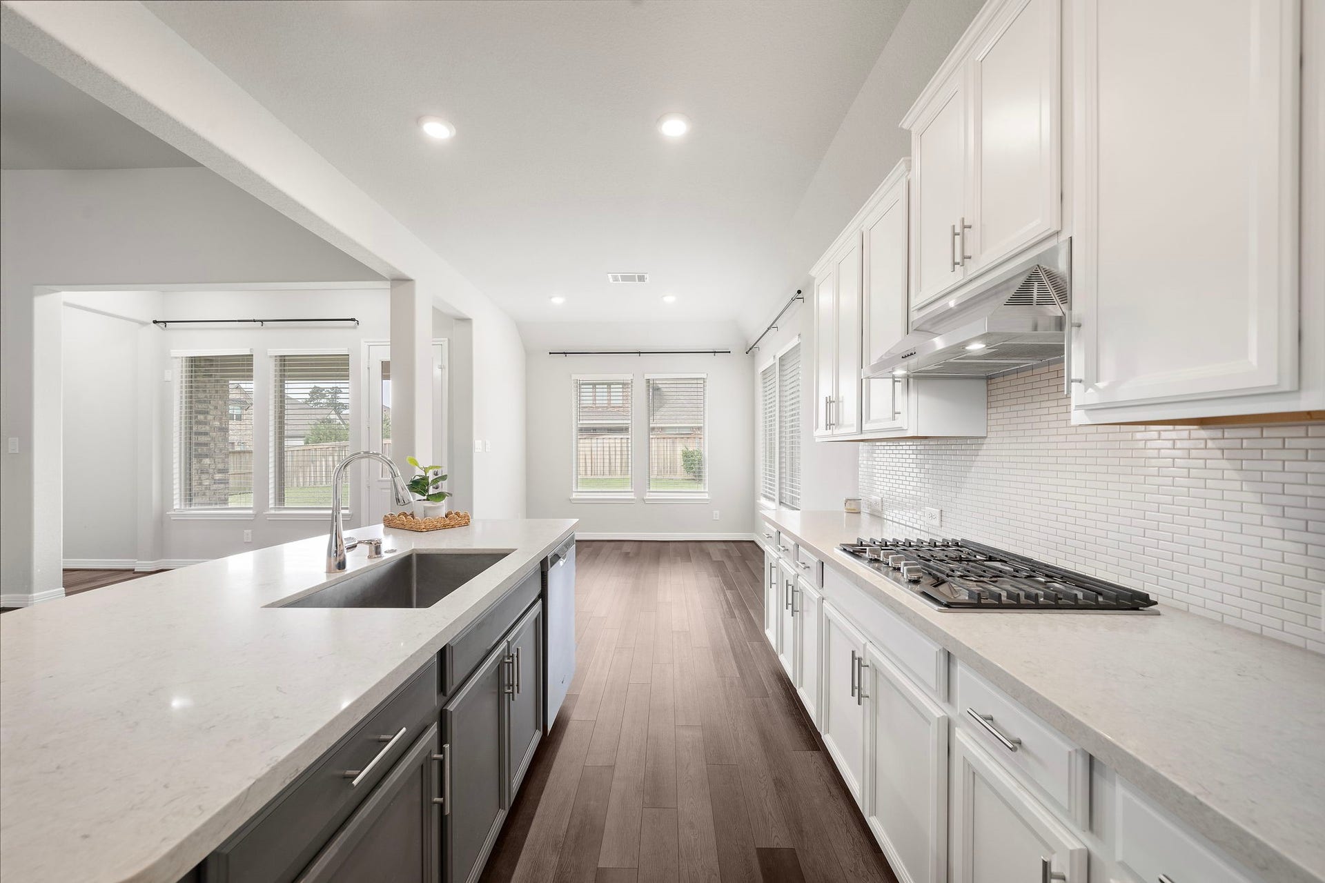 This bright kitchen showcases crisp white cabinetry, sleek countertops, stainless steel appliances, and a spacious island with an undermount sink. A gas cooktop with stainless vent hood, classic tile backsplash, recessed lighting, and abundant natural light create a polished, functional space designed for everyday cooking, gathering, and entertaining.
